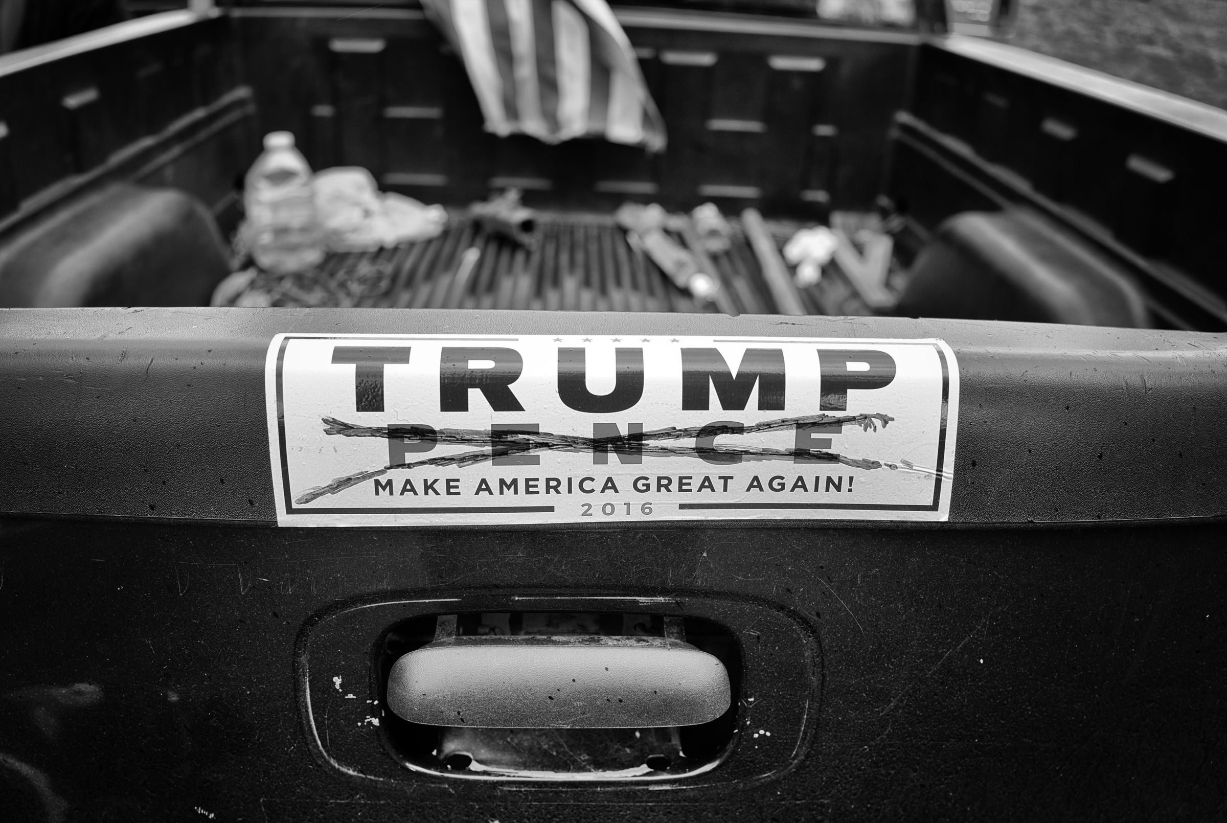 A close-up of a pickup truck tailgate displays a worn "TRUMP PENCE MAKE AMERICA GREAT AGAIN 2016" bumper sticker with Pence's name crossed out with a marker.