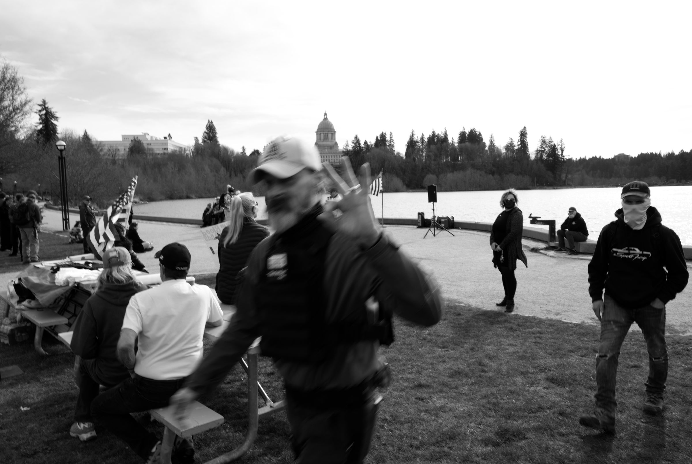 A gathering of people near a body of water, with the Washington State Capitol building visible in the background. A blurry man in the foreground flashes an "OK" hand sign to the camera as he passes. The man is wearing a ball cap and a military-style armored plate carrier. Behind him, a masked man in a ball cap advances menacingly.