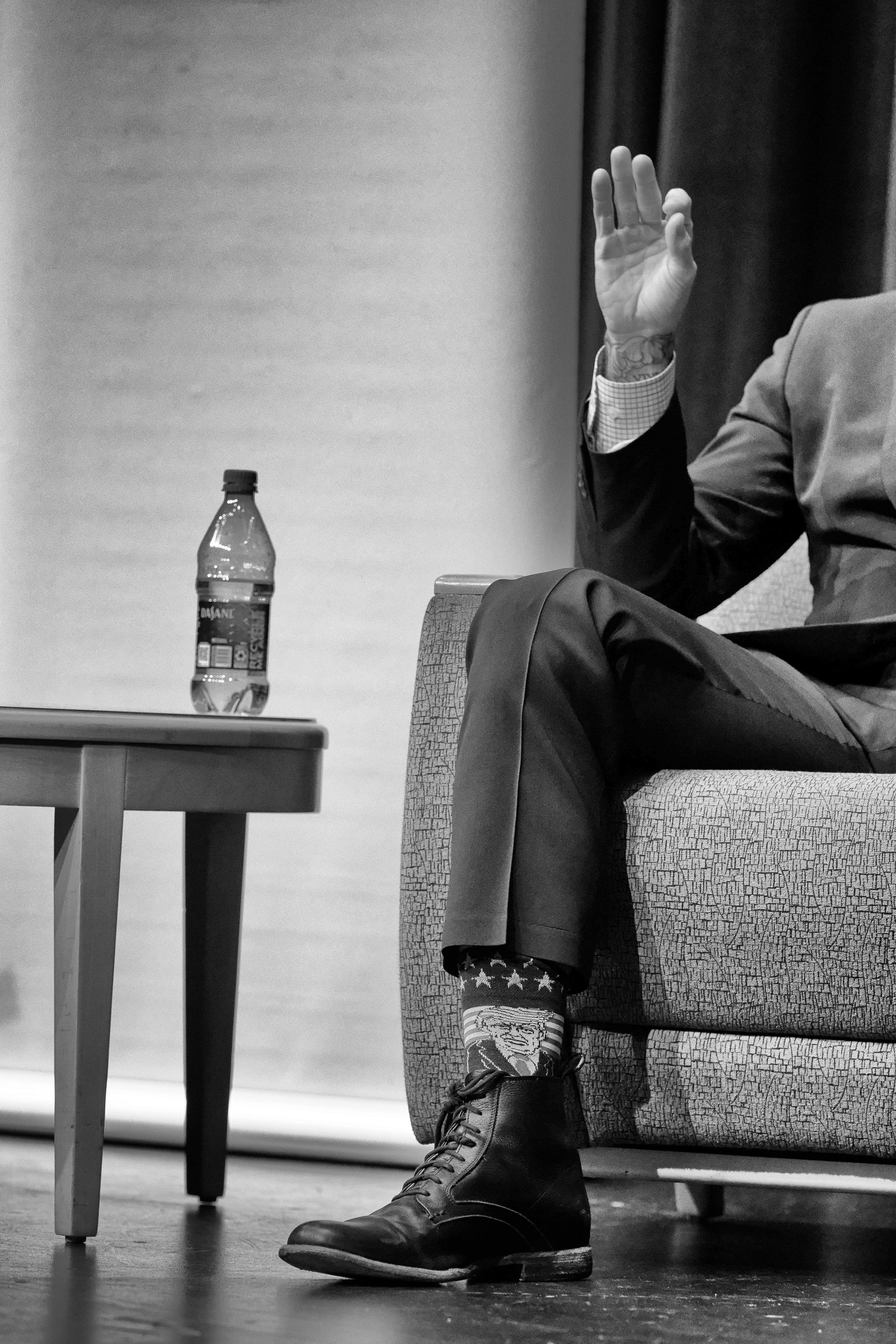 A close-up black and white photo of a person sitting on a stage, wearing a suit with high socks featuring the American flag and an image of Donald Trump. The person's hand is raised in a "zero" or "OK" gesture. A water bottle sits on a small table beside them.