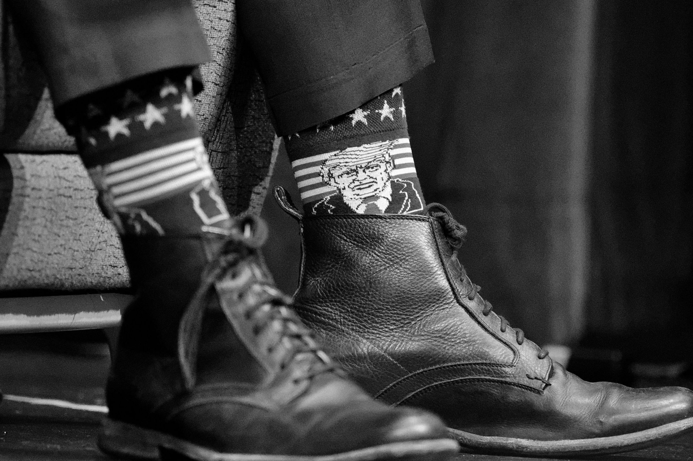 A close-up black and white photo of a man's leather boots shows him wearing high socks featuring stars and stripes and a cartoon depiction of Donald Trump.