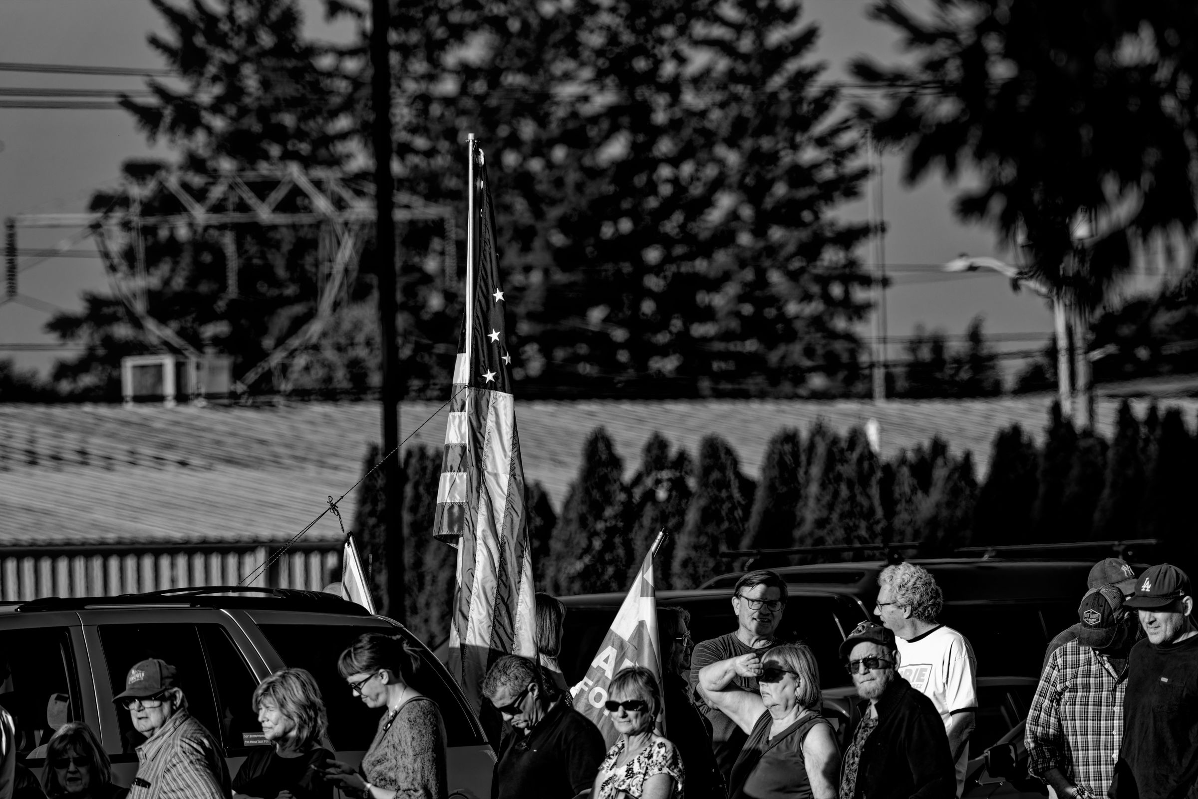 A black and white photo of a group of people standing in front of vehicles and an American flag at a public event, with some individuals looking at the camera while others converse. Trees and utility wires are visible in the background.