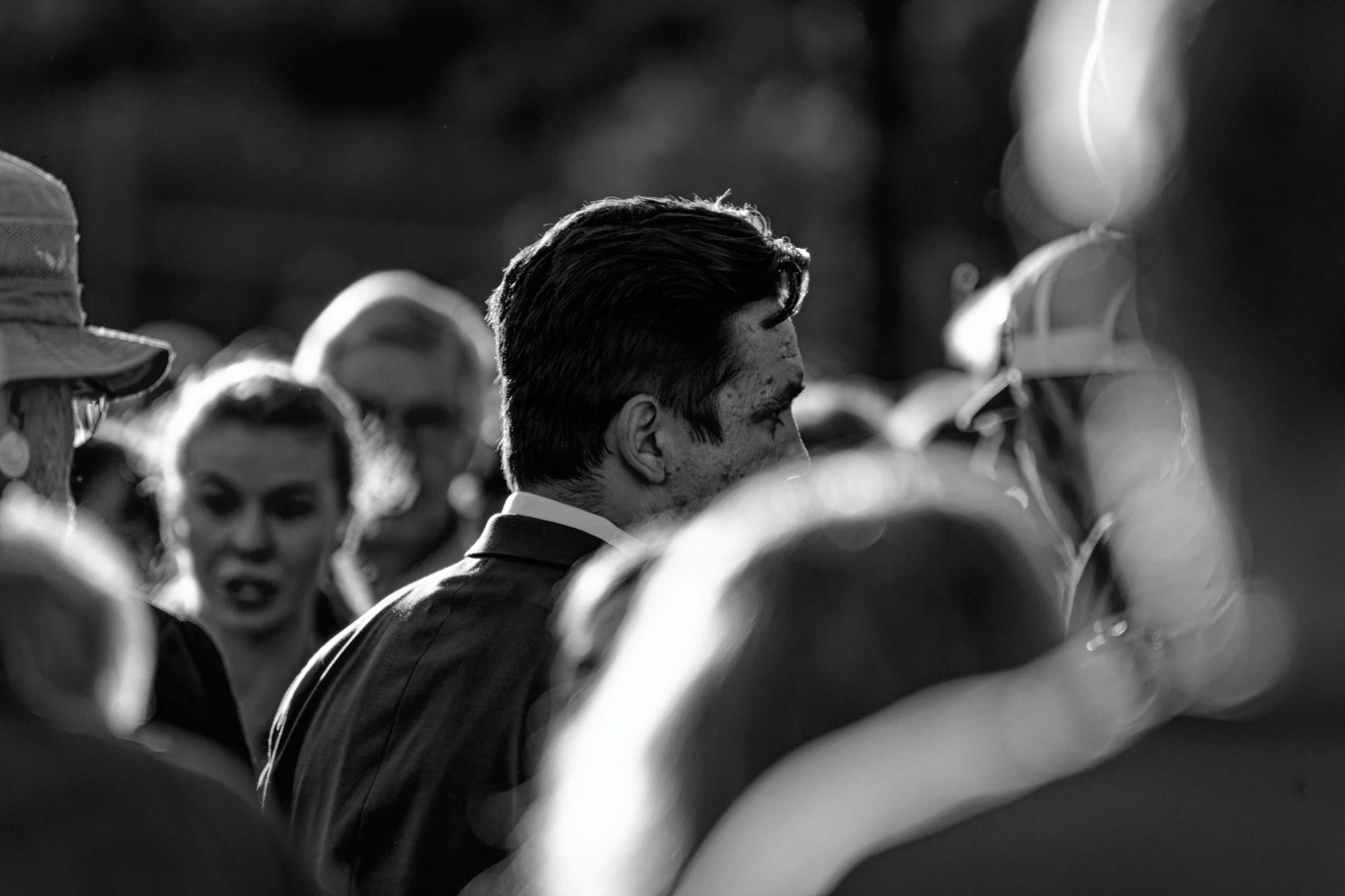 A black and white photo of a man in a suit facing away from the camera in a crowded setting, with people around him engaged in conversation. The sunlight creates dramatic contrasts, emphasizing the back of his head and the intensity of the surrounding scene.