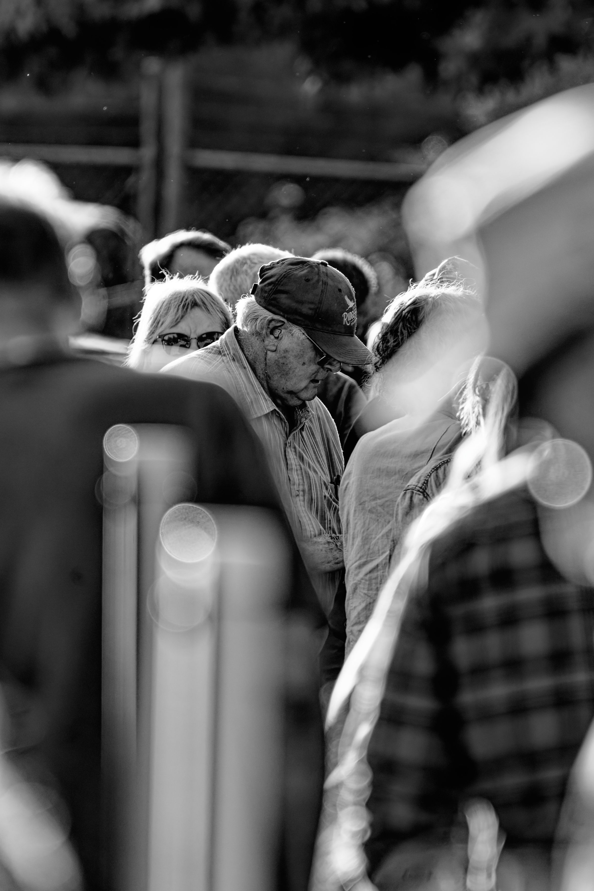 A black and white photo of an elderly man wearing a cap while standing amidst a crowd, his face partially obscured by shadows, with the sunlight highlighting the faces around him. His hat partially reads "Ridgefield" and depicts a bird in flight.