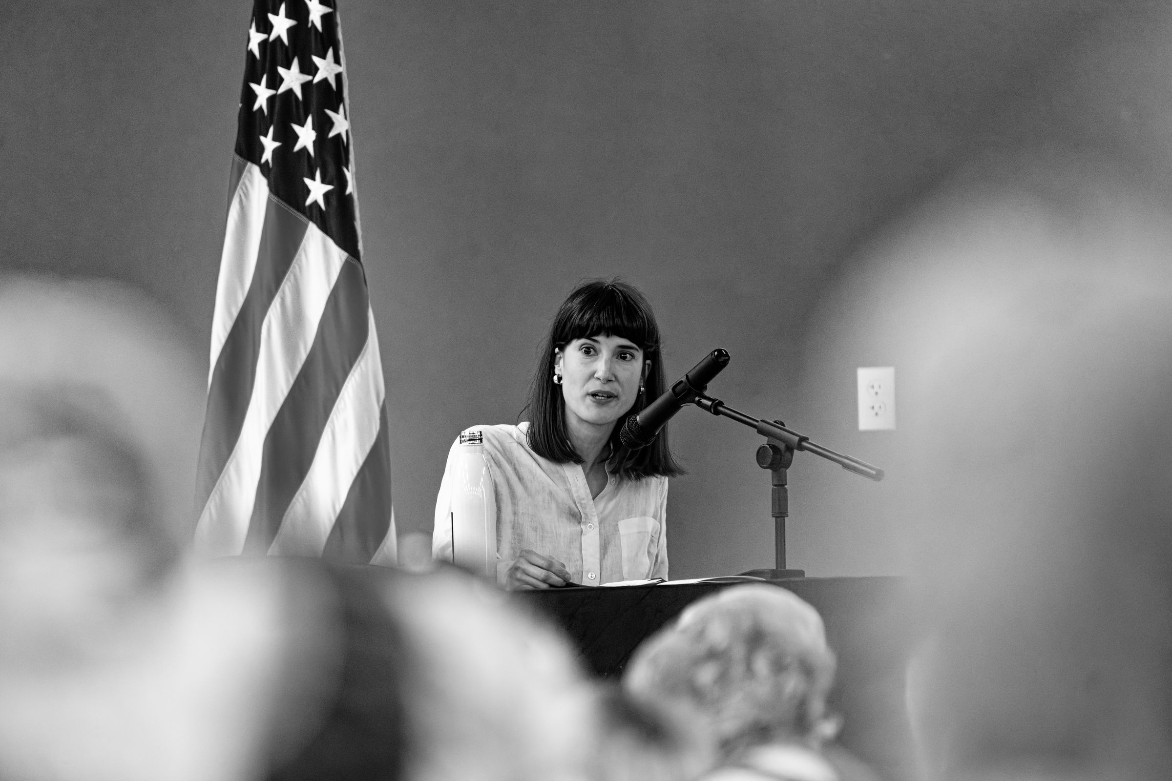 A black and white photo of Marie Gluesenkamp Perez wearing a light formal button-up shirt, seated at a table with a microphone in front of her, speaking earnestly to a crowded auditorium with the American flag standing prominently beside her.