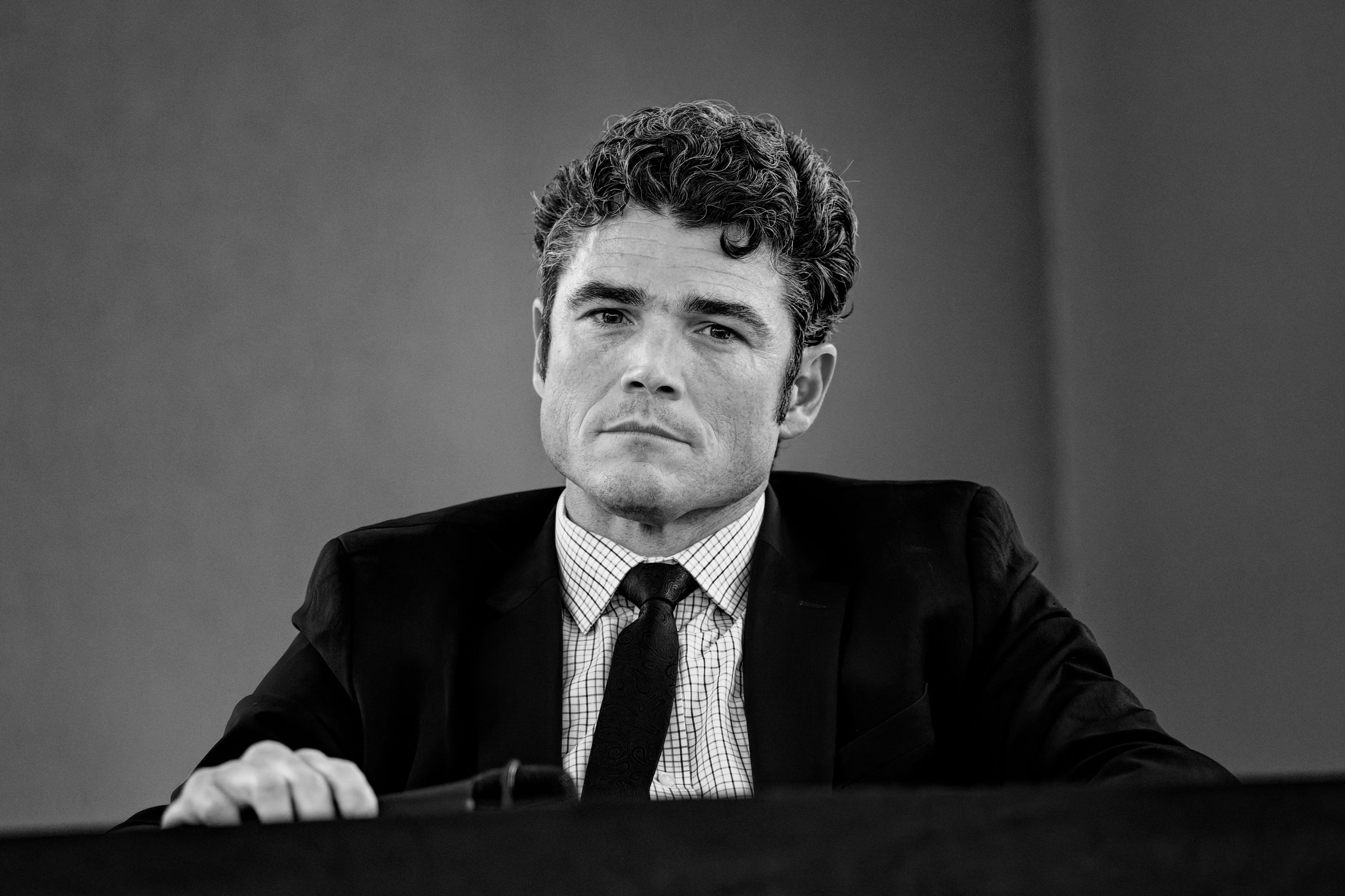 A close-up of Joe Kent in a suit and tie, looking solemnly at the camera while holding a microphone at a podium.