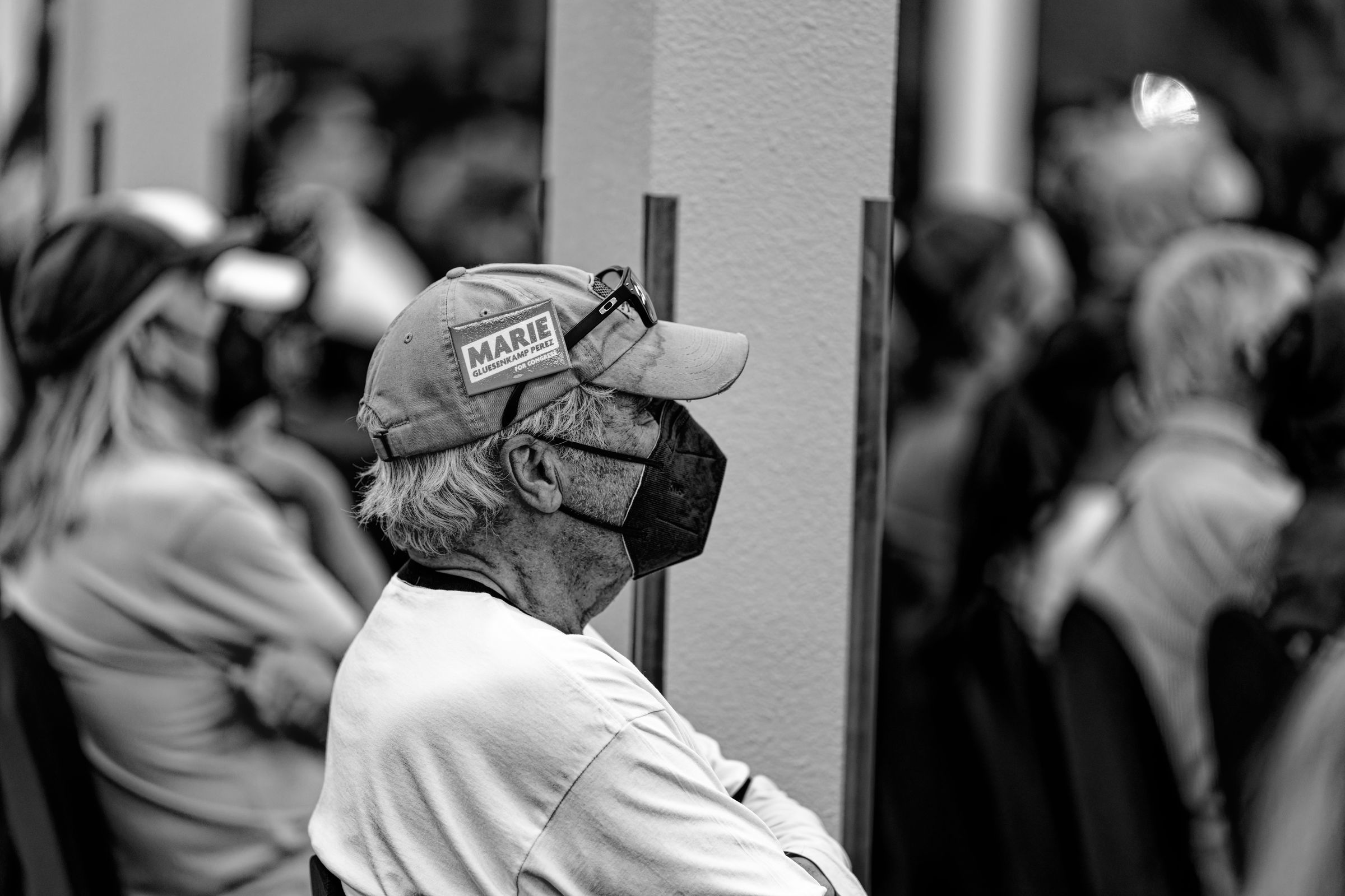 A black and white photo of an elderly man with a face mask and a hat with a "Marie Gluesenkamp Perez" pin on it, sitting with his arms crossed in a crowded auditorium.