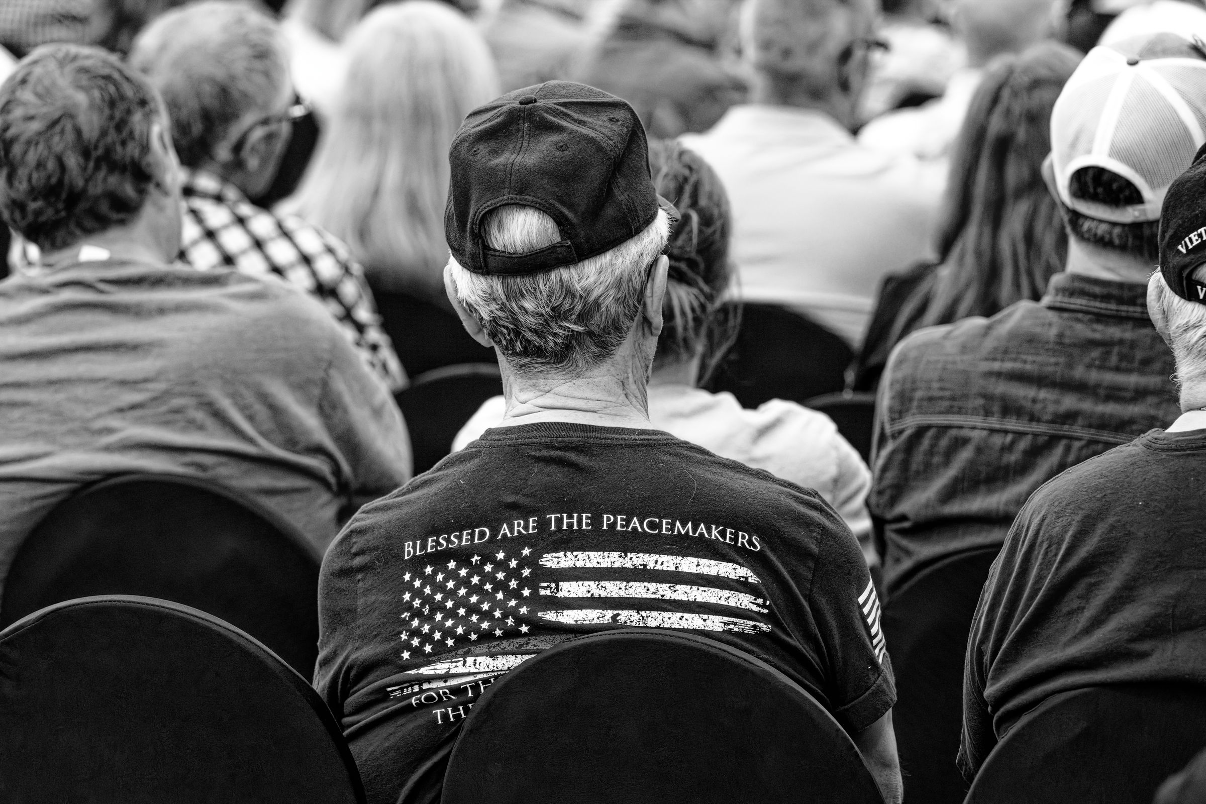 A black and white photo of a man with short gray hair and a cap, facing away from the camera. He wears a black shirt with an American flag and the words "Blessed Are The Peacemakers" on the back, sitting among other attendees in a crowded auditorium.