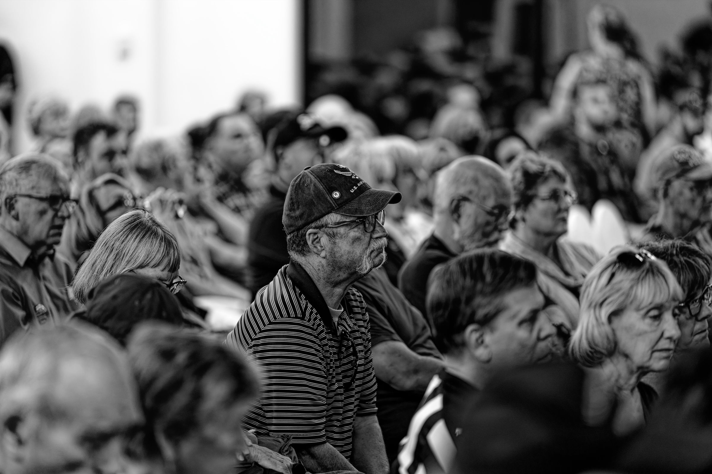 A black and white photograph of a man wearing glasses, a striped shirt, and a U.S. Air Force ball cap while sitting attentively in a crowded auditorium.