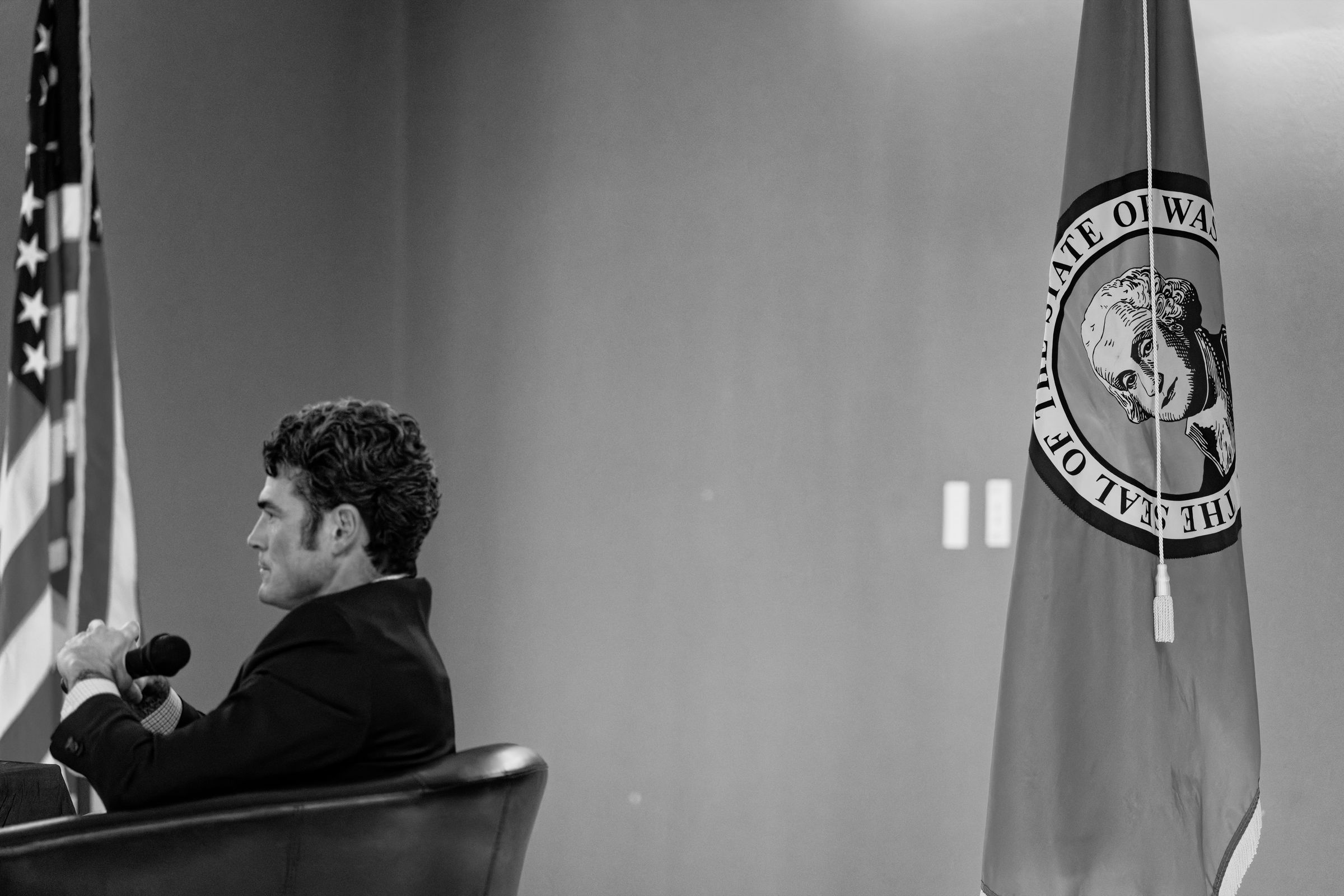 A black and white photo of Joe Kent in a suit, seated at a table with a microphone, facing away from the camera. A U.S. flag stands in the background to his front, and the Washington state flag is focused in the foreground to his back. On the Washington state flag, George Washington's eyes appear to look at the camera.