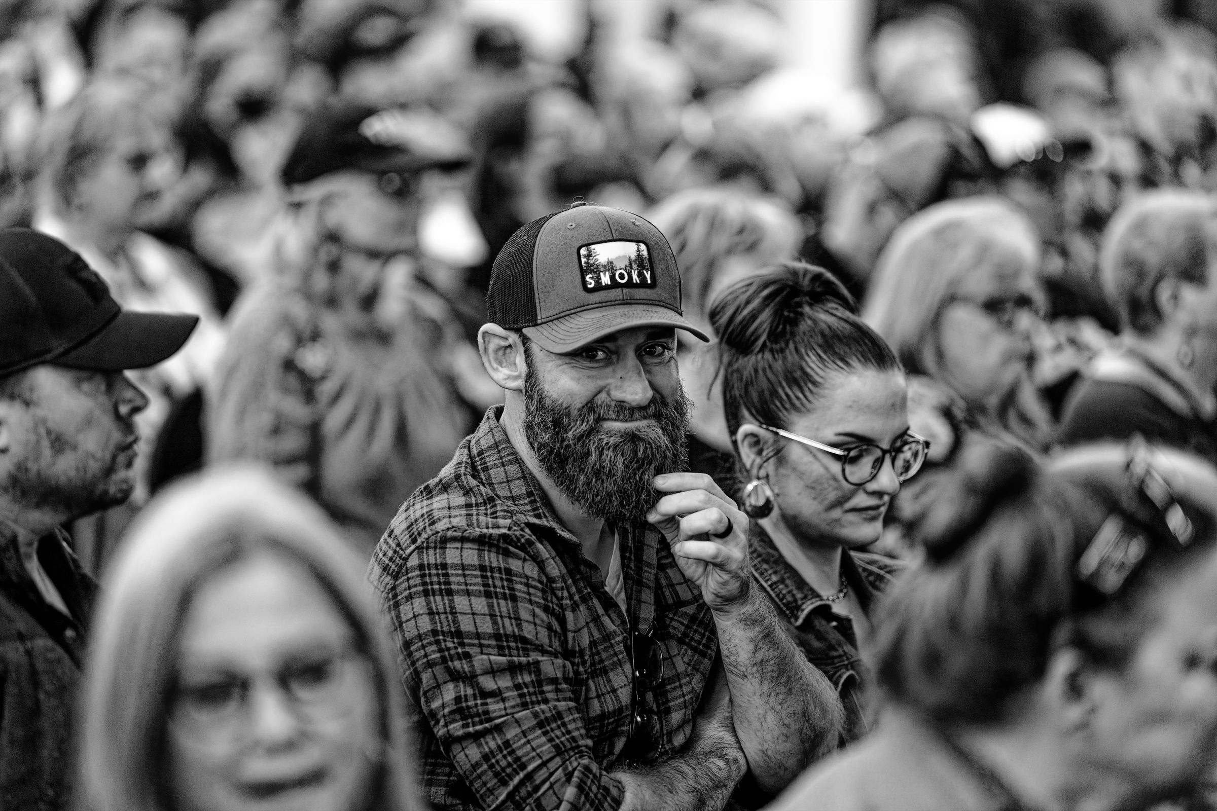 A close-up black and white photograph of a bearded man in a plaid shirt and baseball cap, surrounded by other people in a crowded auditorium. The man is smiling at the camera while stroking his beard.