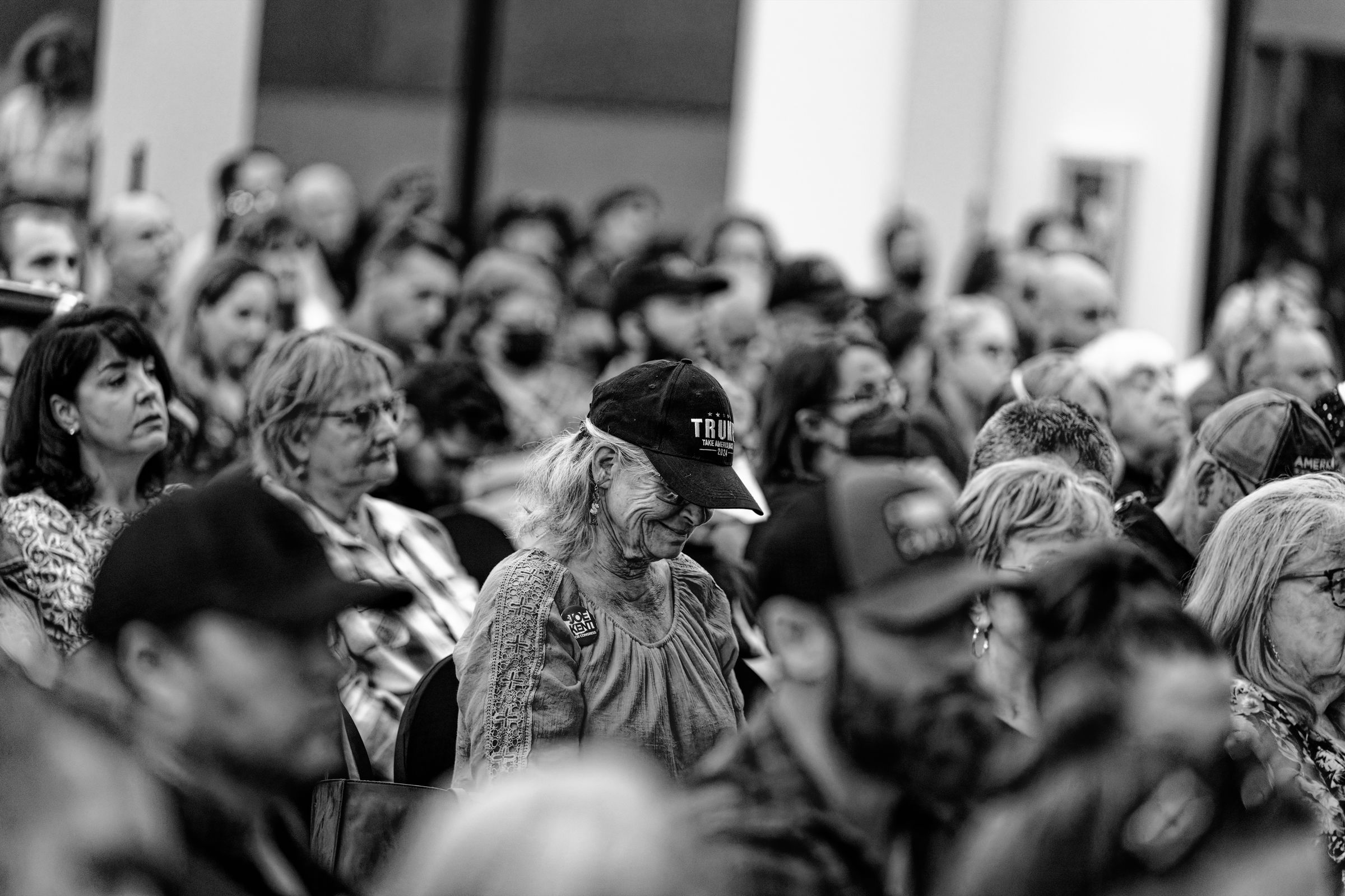 A black and white image of a diverse crowd at a political event. In the foreground, an older woman wearing a TRUMP campaign hat visibly smiles among the seated attendees.