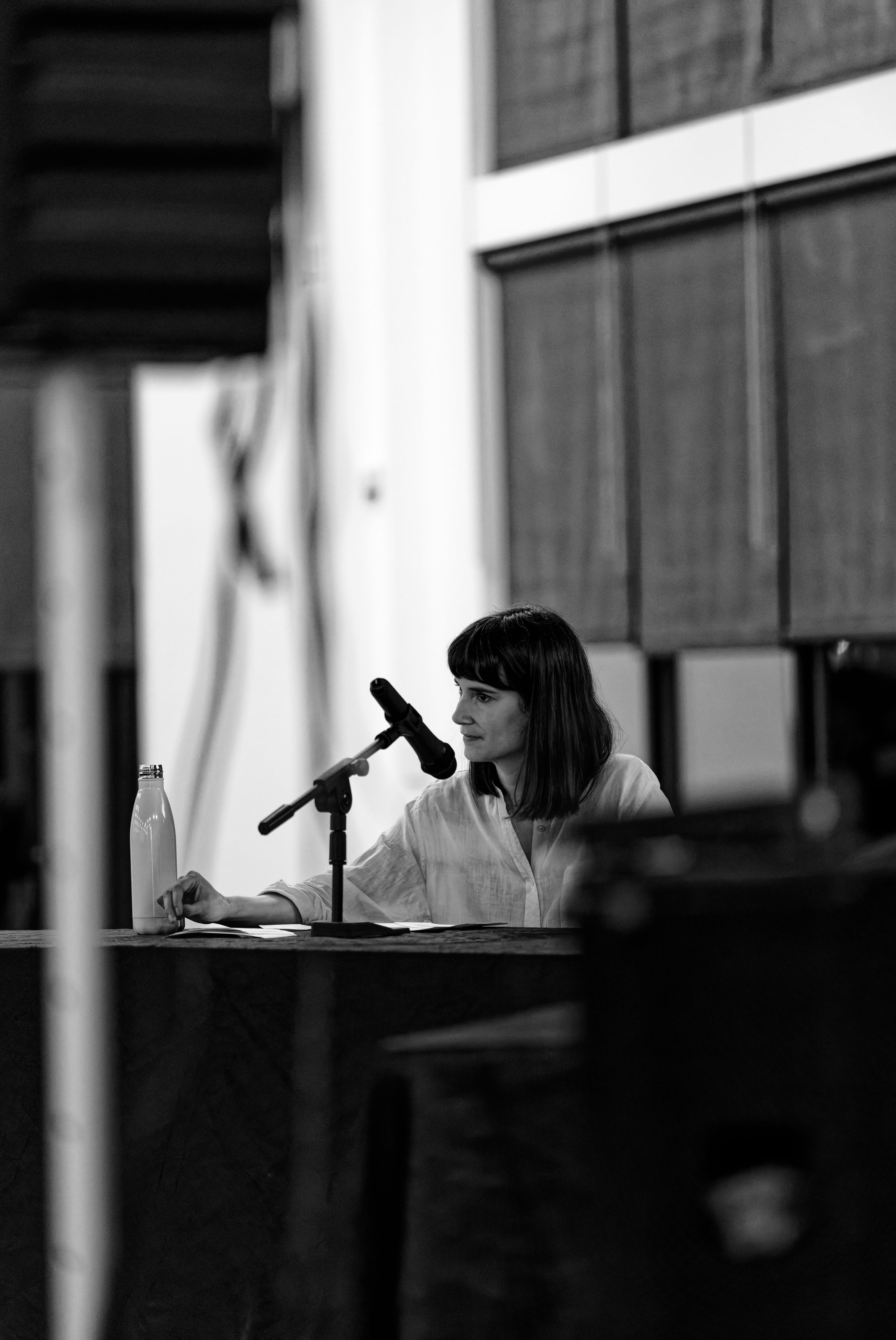 A black and white photograph of Marie Gluesenkamp Perez speaking at a podium, addressing an audience in a large auditorium.