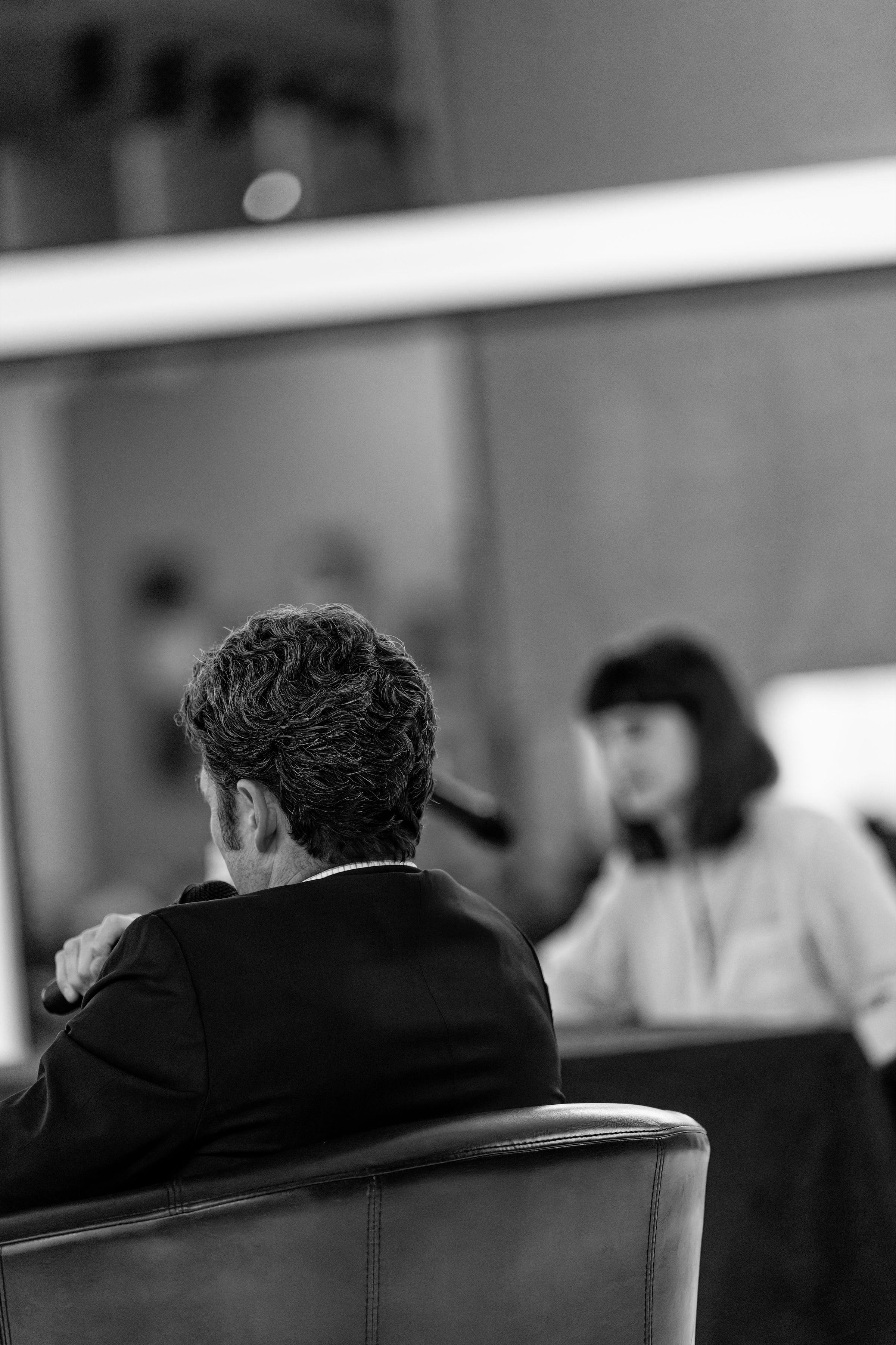 A black and white photo of Joe Kent seated at a table, facing Marie Gluesenkamp Perez as he speaks into a microphone.