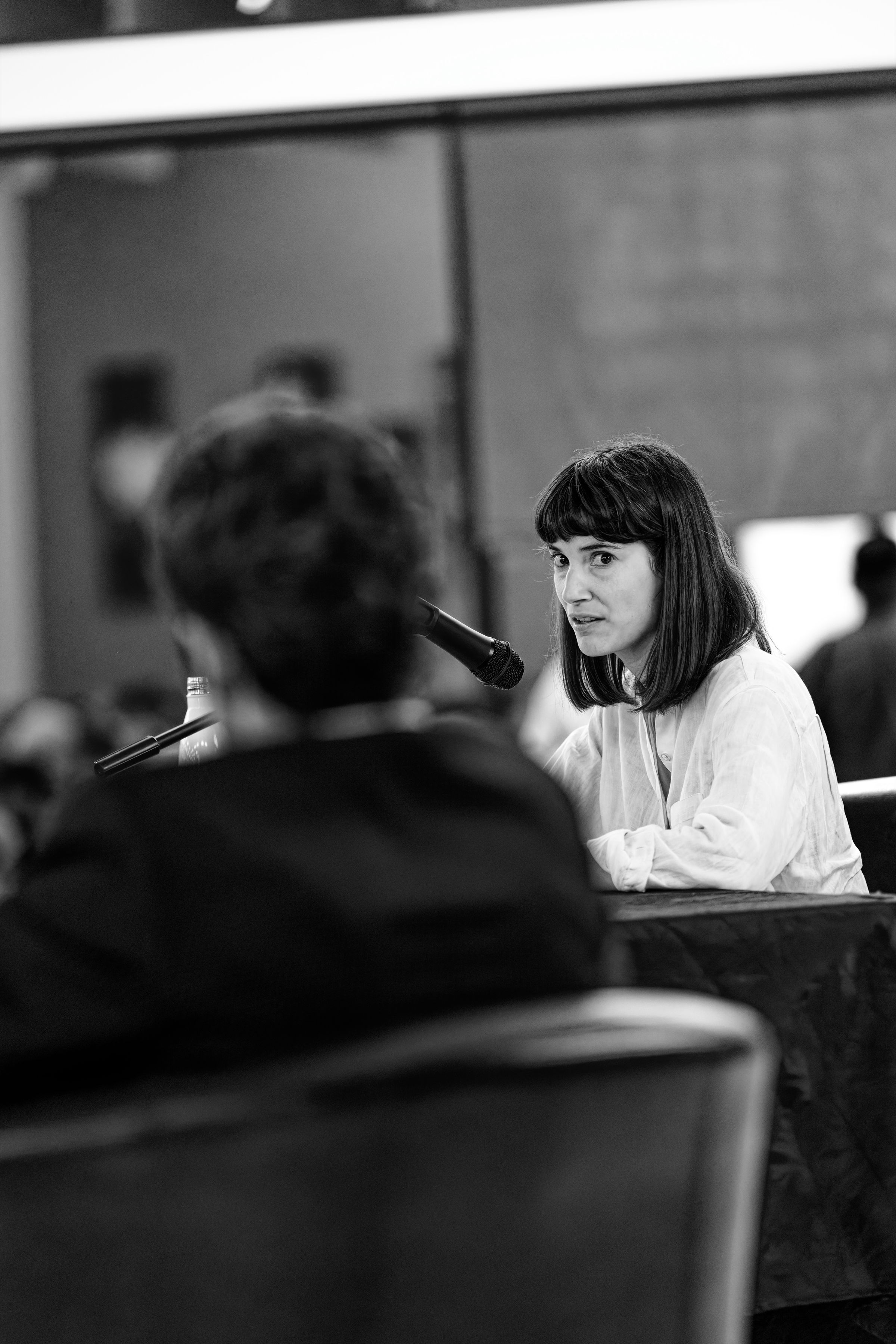 A black and white photograph of two seated speakers at a podium during a political debate. Marie Gluesenkamp Perez speaks into a microphone while looking intensely at Joe Kent in the foreground.