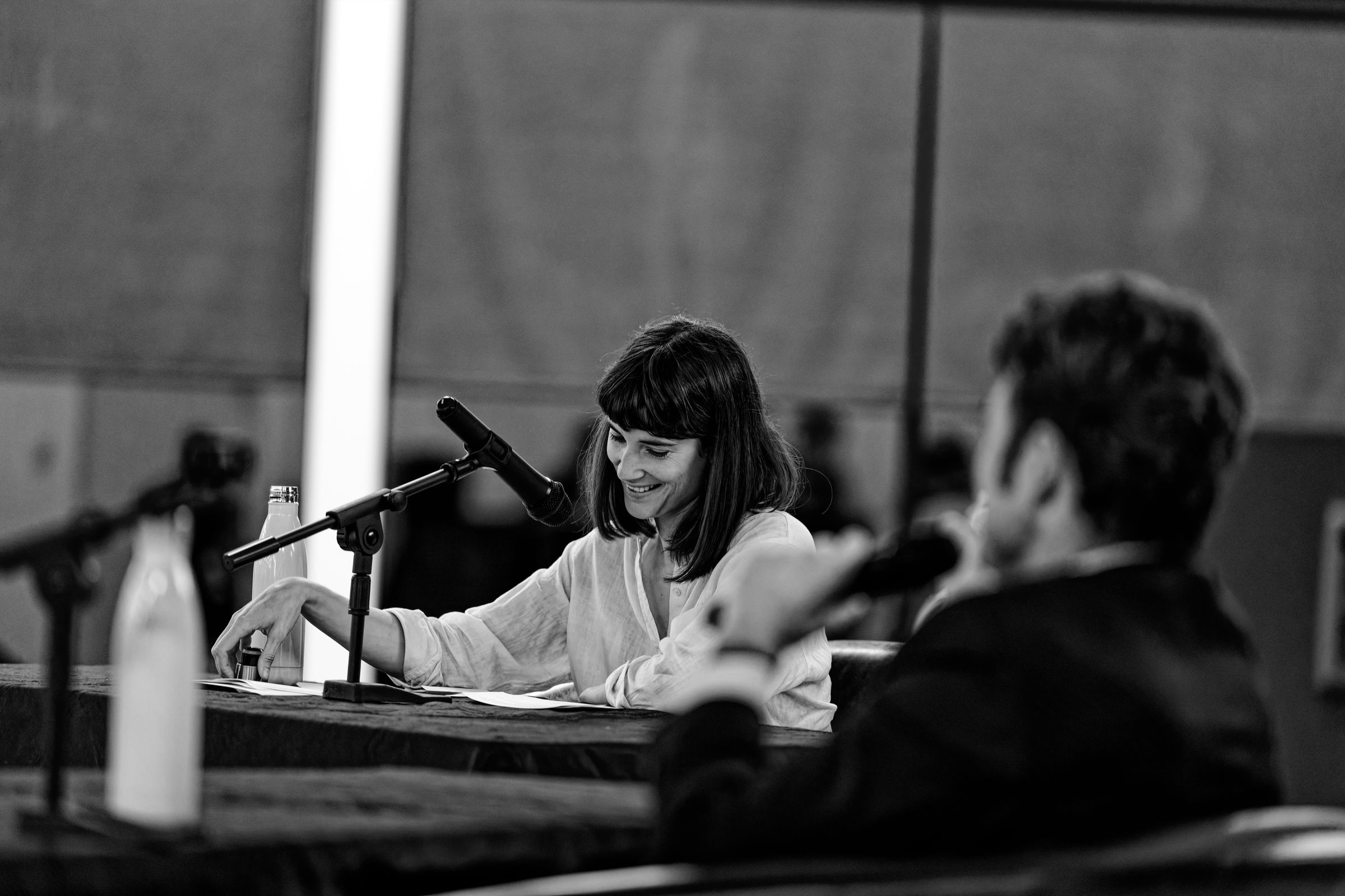 A black and white photograph of two speakers at a podium during a political debate. Marie Gluesenkamp Perez smiles as Joe Kent speaks into a microphone in the foreground.