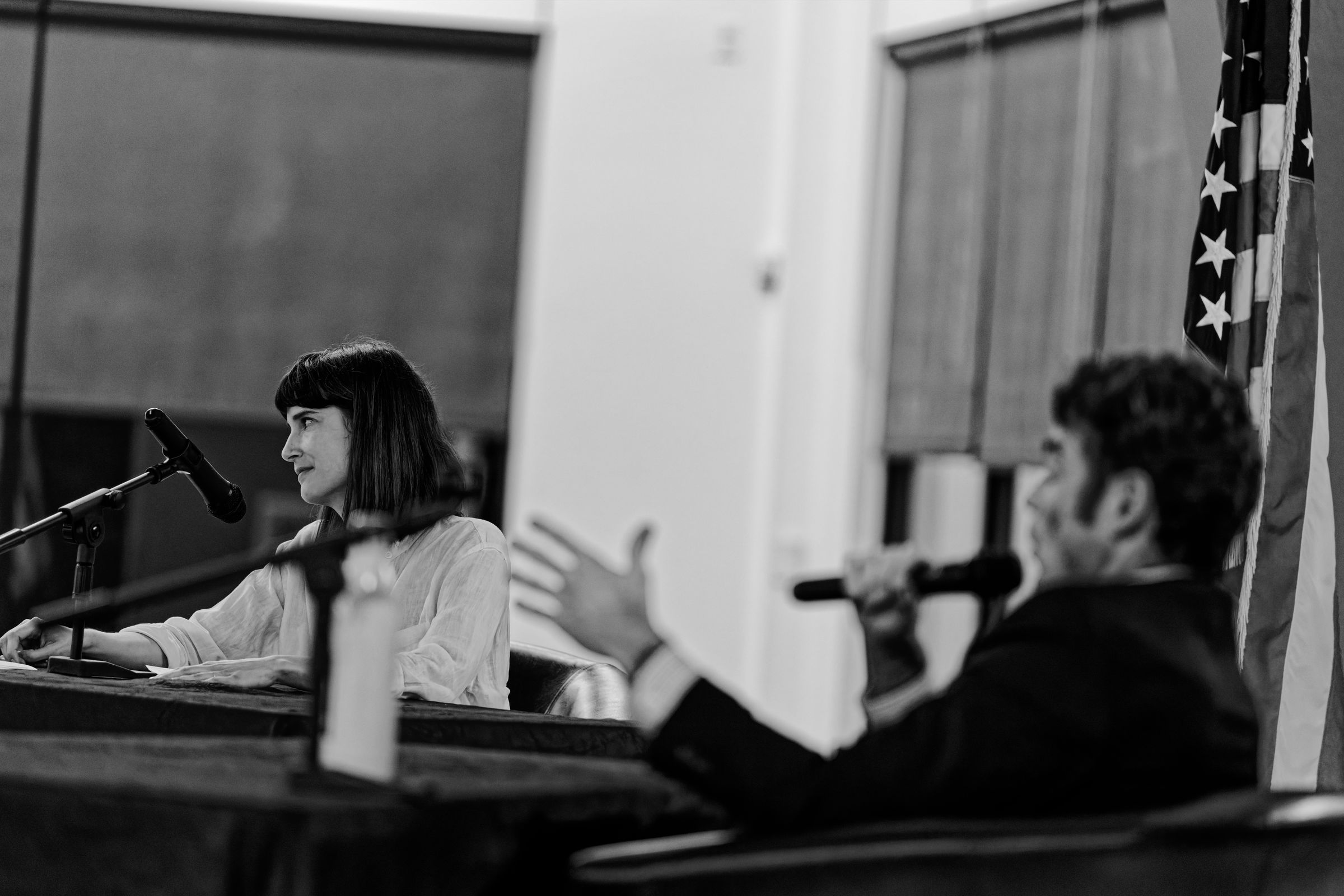 A black and white photograph of two speakers at a podium during a political debate. Marie Gluesenkamp Perez looks amusedly towards the audience while Joe Kent gesticulates wildly in the foreground. An American flag is visible in the background.