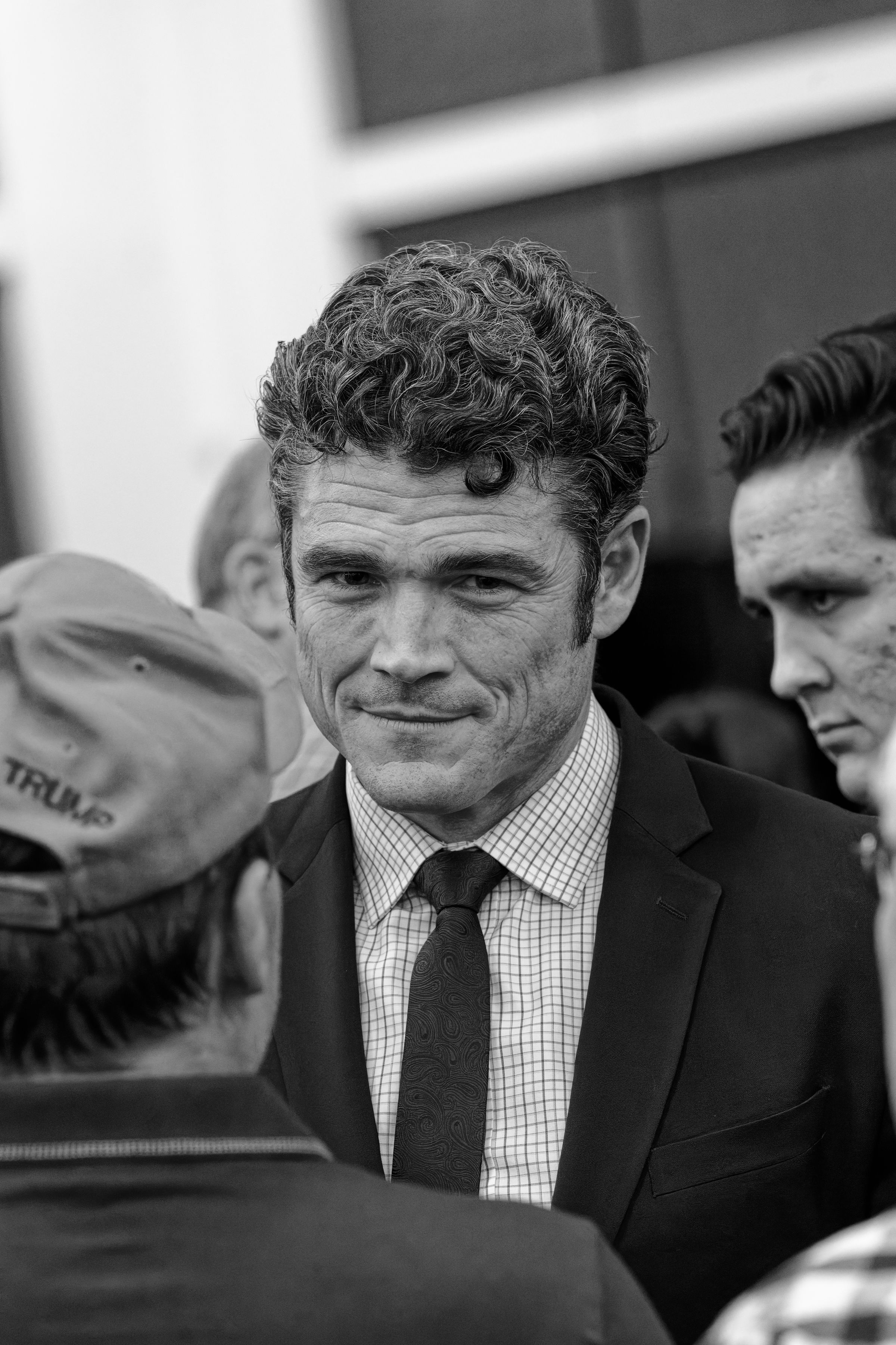 A black and white close-up portrait of Joe Kent wearing a suit and tie. He's surrounded by supporters, one wearing a Trump campaign hat partially visible in the foreground.