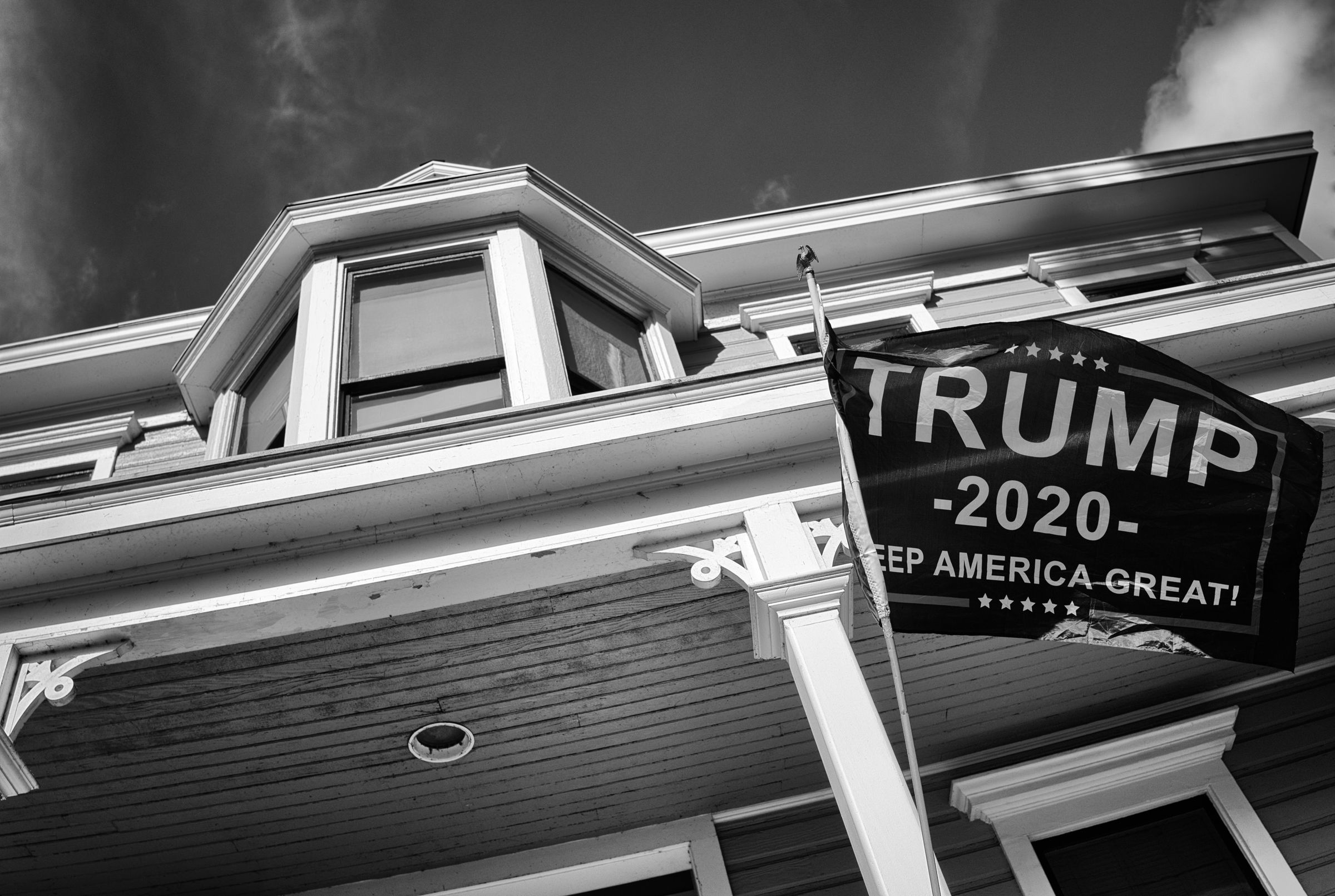 A low-angle black and white photograph of a Victorian-style house exterior. A Trump 2020 campaign flag is prominently displayed from a support column.