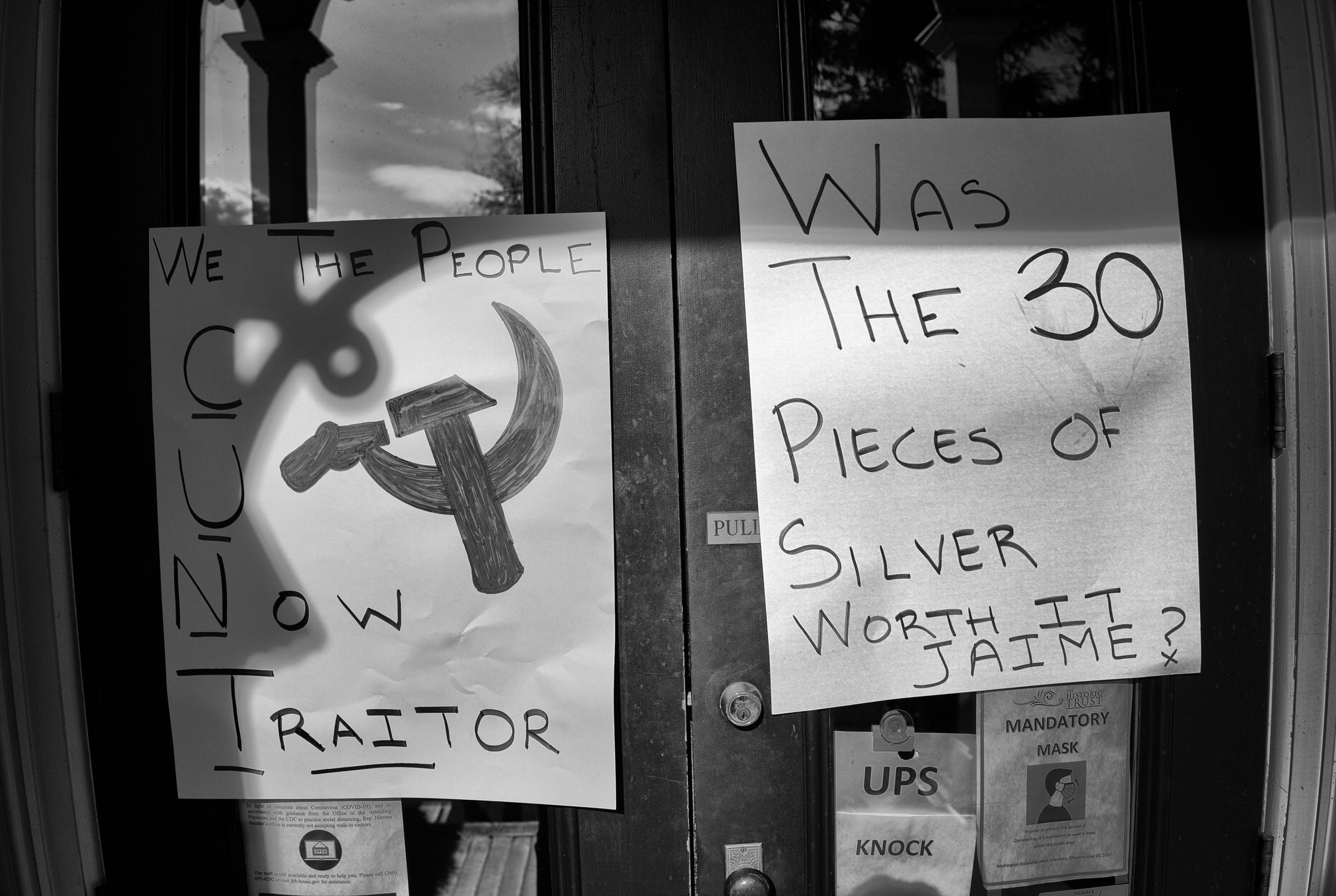 A black and white photo of two handwritten protest signs on a glass door. One sign displays a hammer and sickle drawing with the words "We The People C U Now Traitor", with the letters C, U, N, and T underlined. The other sign reads "Was The 30 Pieces Of Silver Worth It Jaime?"