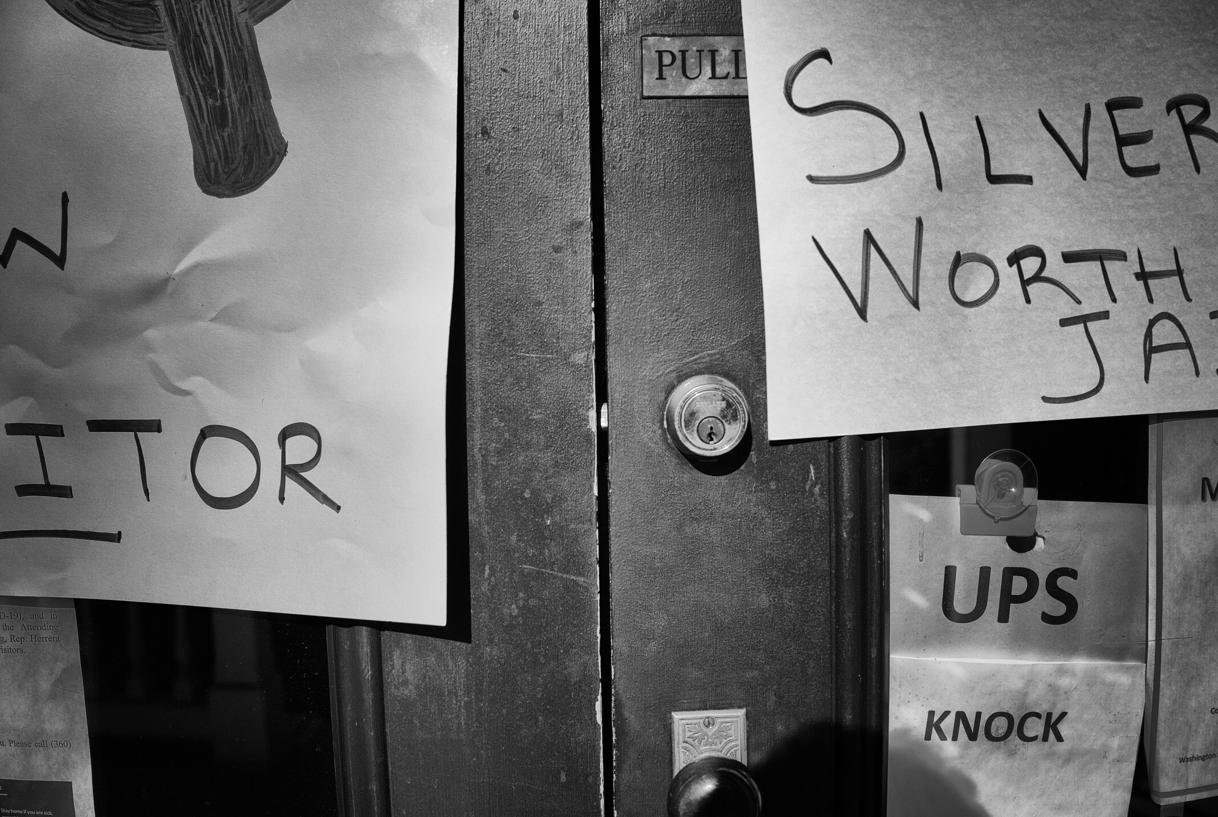 A close-up black and white image of a wooden door with handwritten signs attached. The signs contain partial words and phrases, with "Silver Worth" and "ITOR" visible. The door's locked deadbolt is focused in the center of the frame.