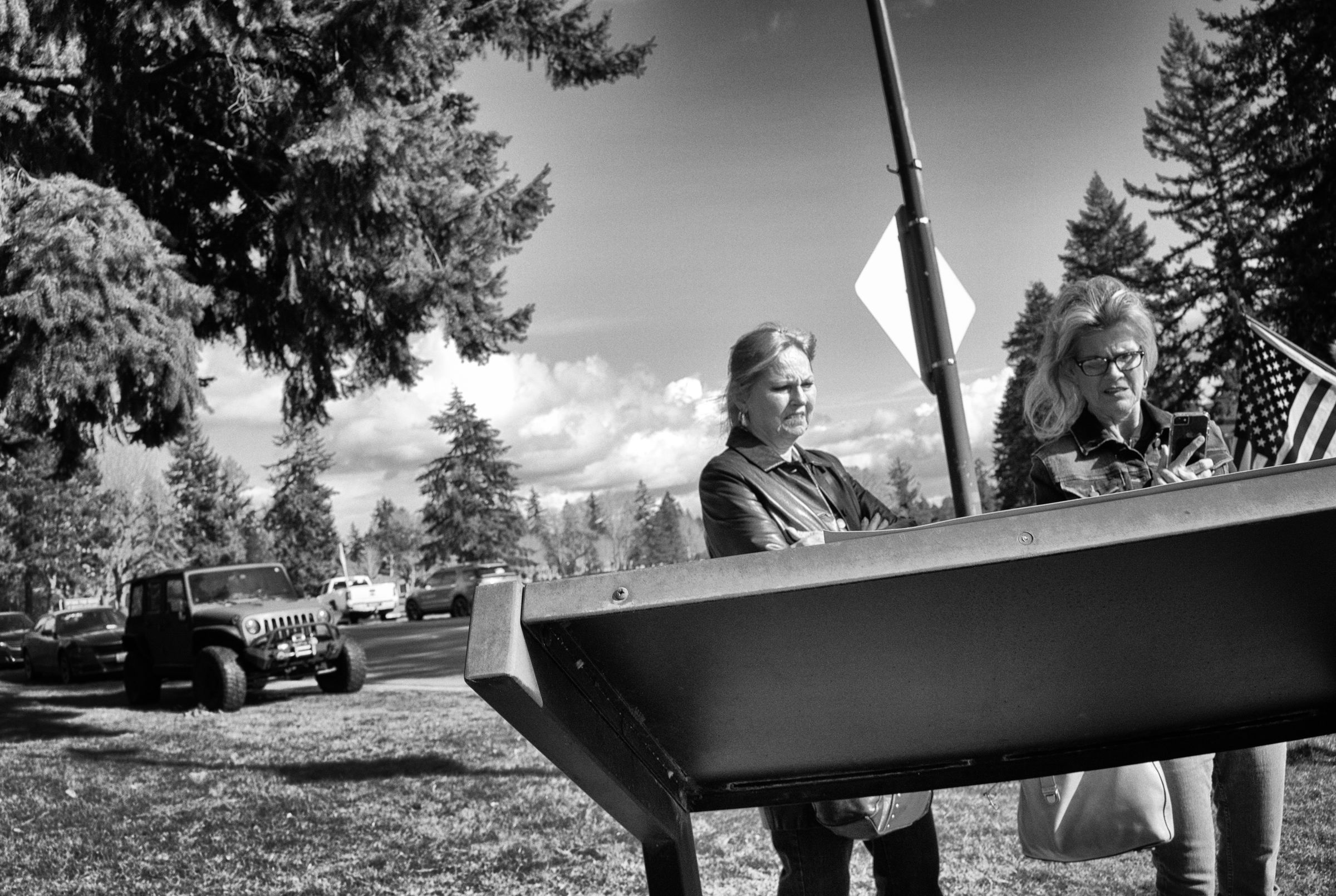 A black and white photograph of two women standing in front of an informational plaque in a park-like setting. An American flag is partially visible behind them, and cars are parked in the background. The women's faces show perplexity and disgust as they read the plaque in front of them.