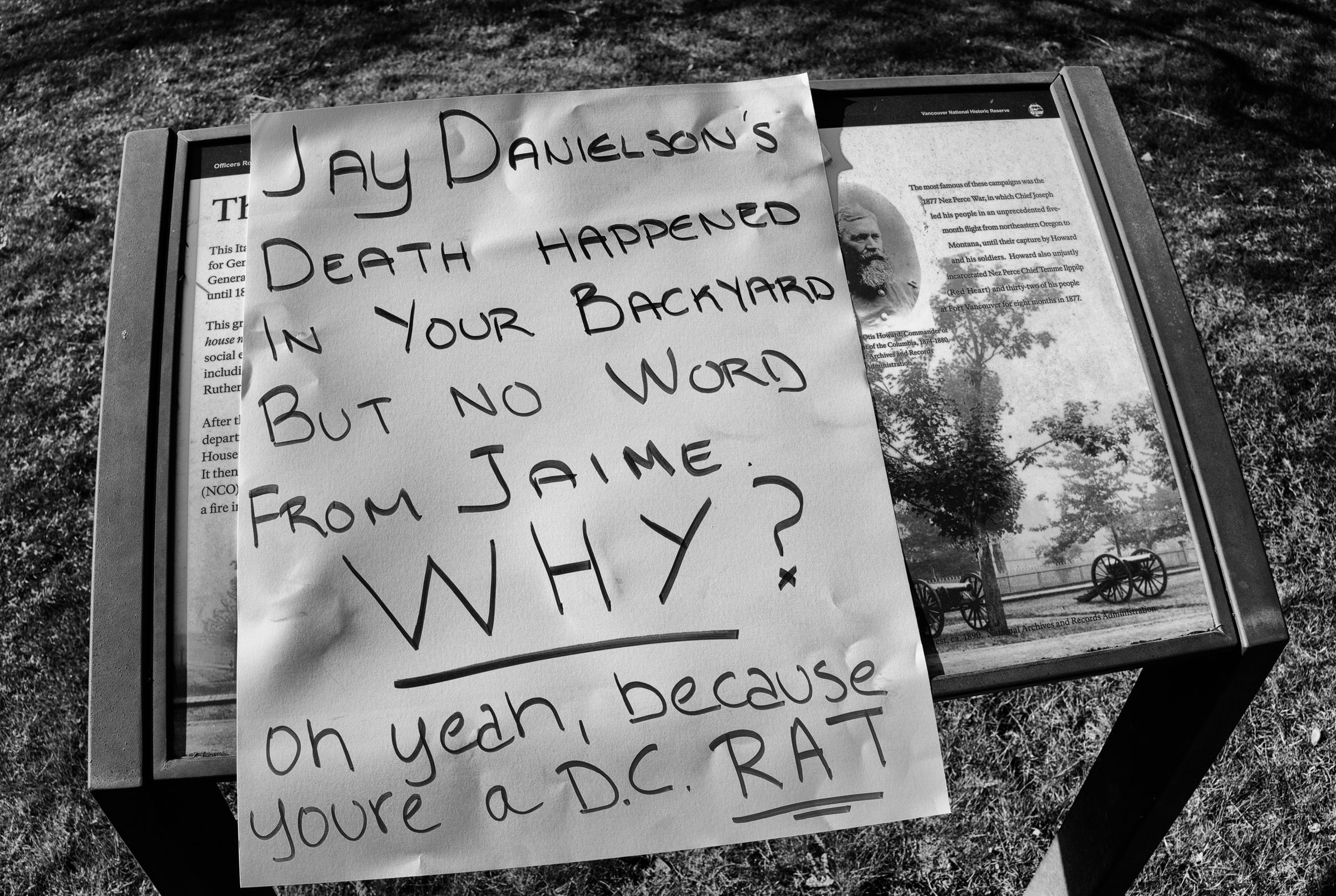 A black and white photo of a handwritten protest sign placed on top of an informational plaque. The sign reads "Jay Danielson's Death Happened In Your Backyard But No Word From Jaime. Why? oh yeah, because youre a D.C. RAT". The word "RAT" is underlined twice.