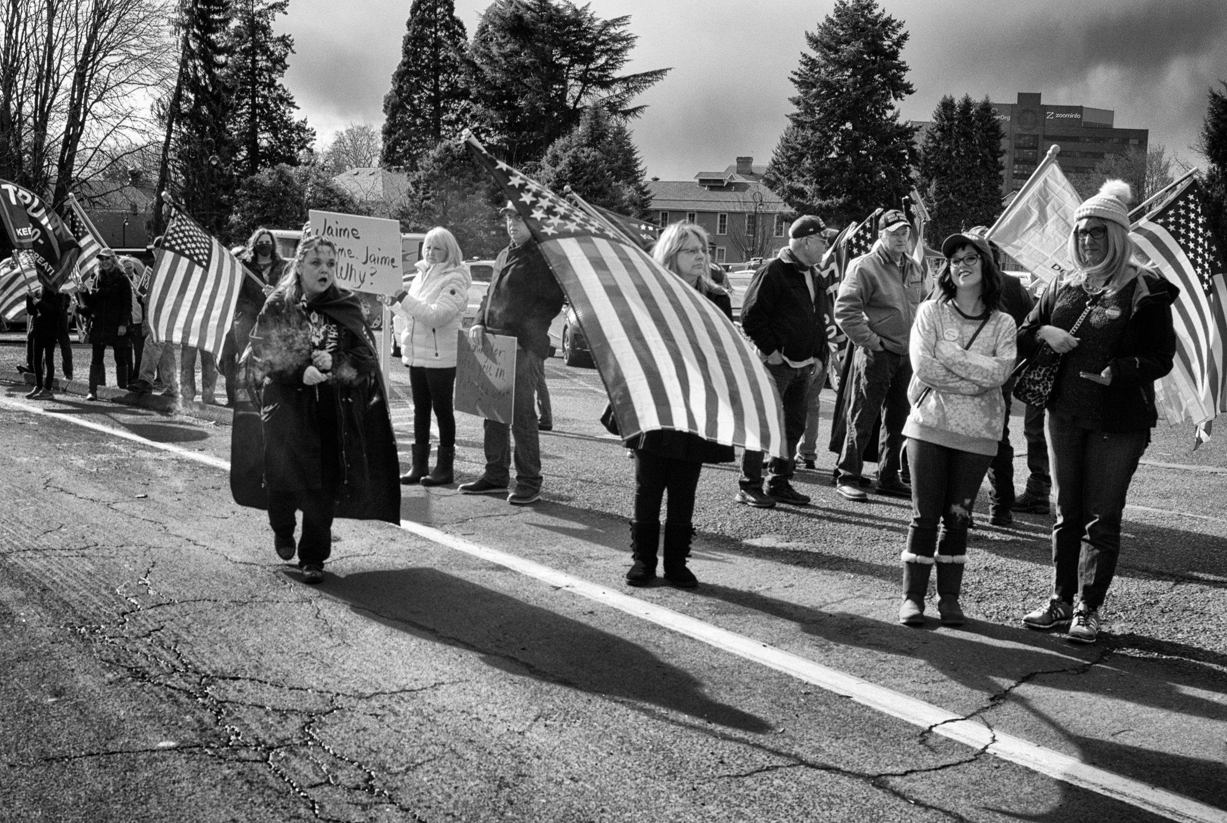 A black and white photograph of a political demonstration. Participants carry American flags and signs while standing along a roadside. Trees and buildings are visible in the background. A partially obscured handmade sign reads "Jaime Why?" Two attendees smile derisively towards the camera.