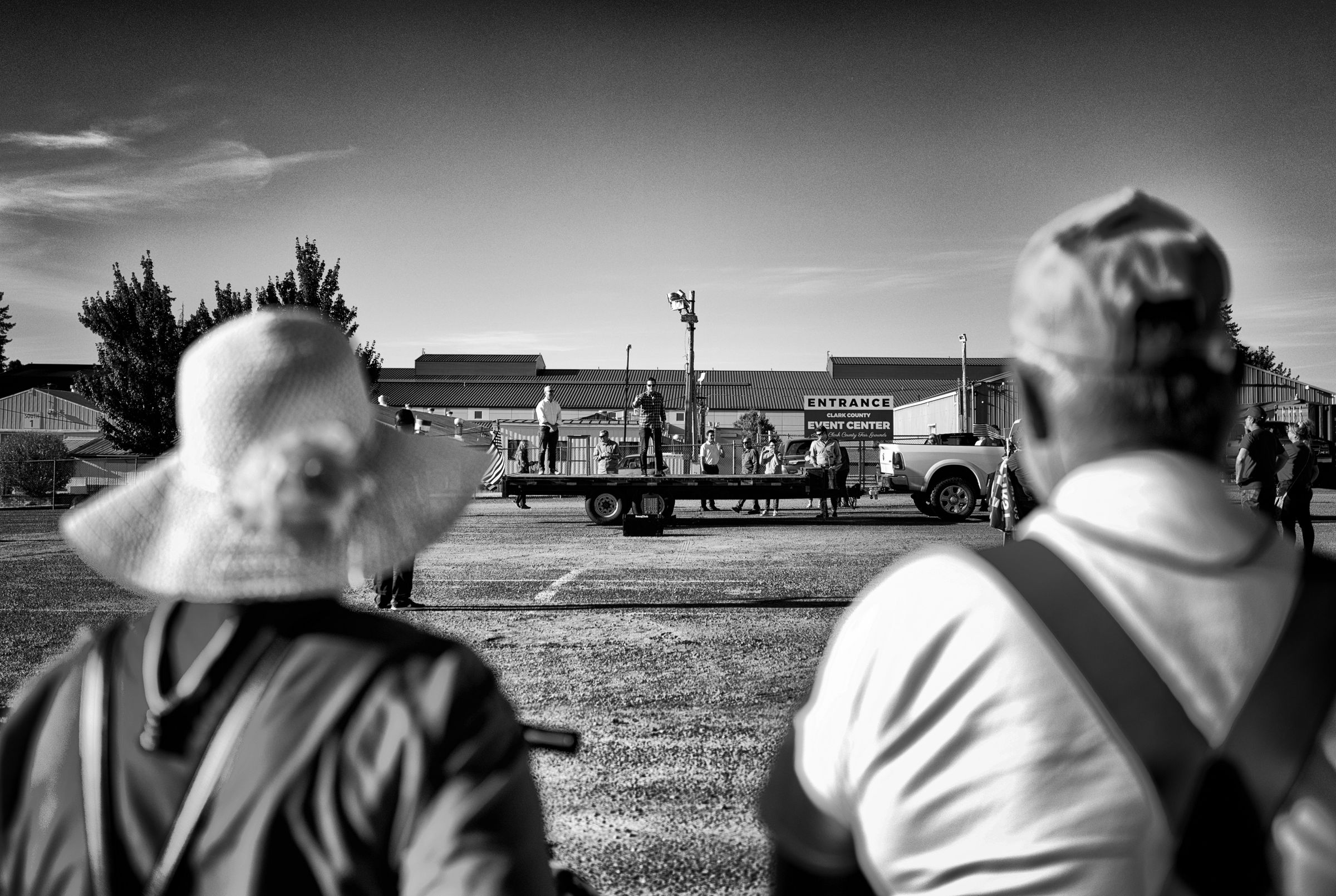 A black and white photo of a rural event center with a flatbed trailer serving as a stage. Two attendees in the foreground observe the scene, one wearing a wide-brimmed sun hat. Florida congressman Matt Gaetz stands on the flatbed trailer in the distance, flanked by campaign staff, speaking while pointing towards the audience.