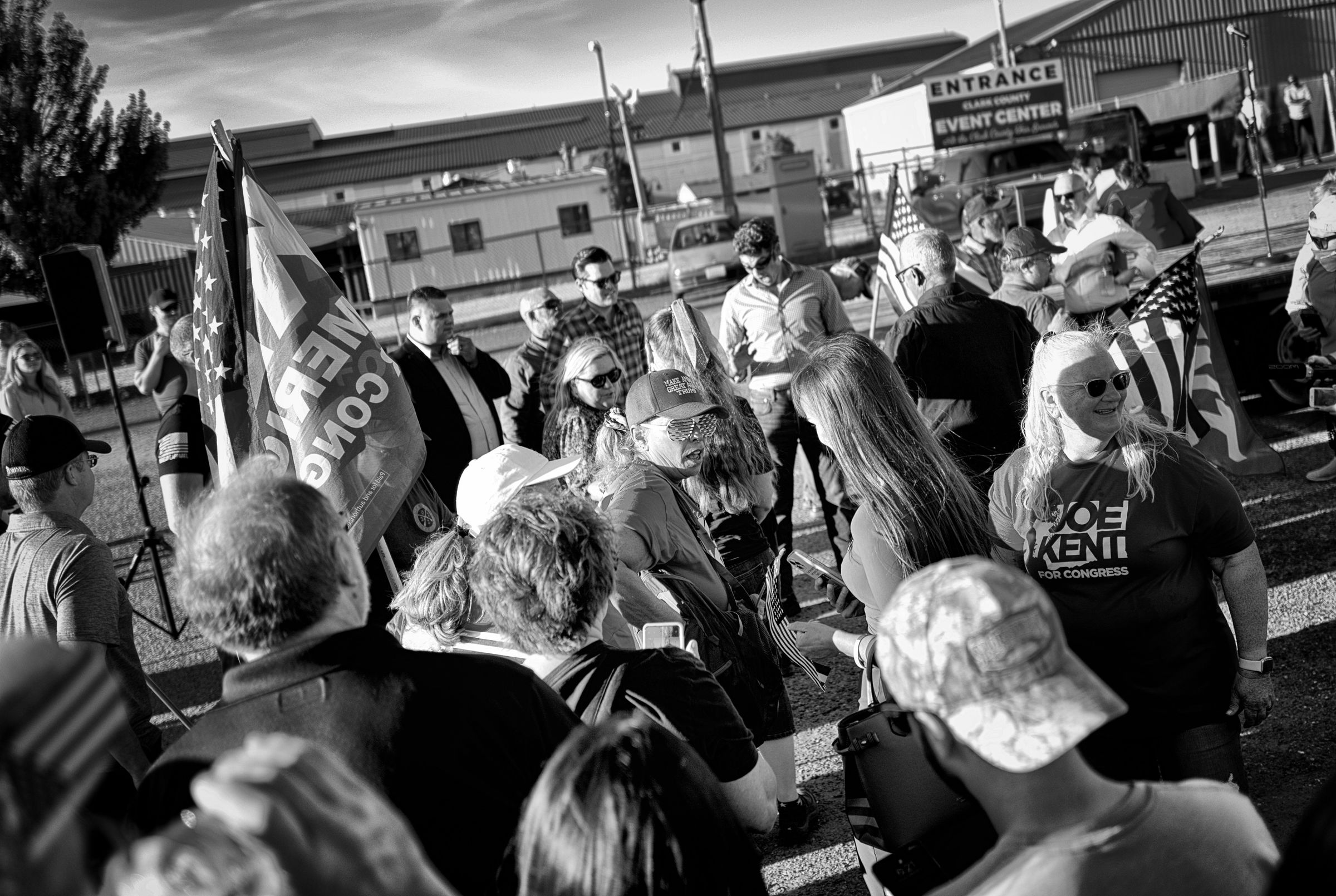 A black and white photo of a crowded political rally showing supporters holding flags and wearing "JOE KENT FOR CONGRESS" campaign merchandise while standing in a loosely organized line. Joe Kent stands at the front of the line, greeting supporters. Florida congressman Matt Gaetz stands to Kent's right, with security personnel surrounding them.