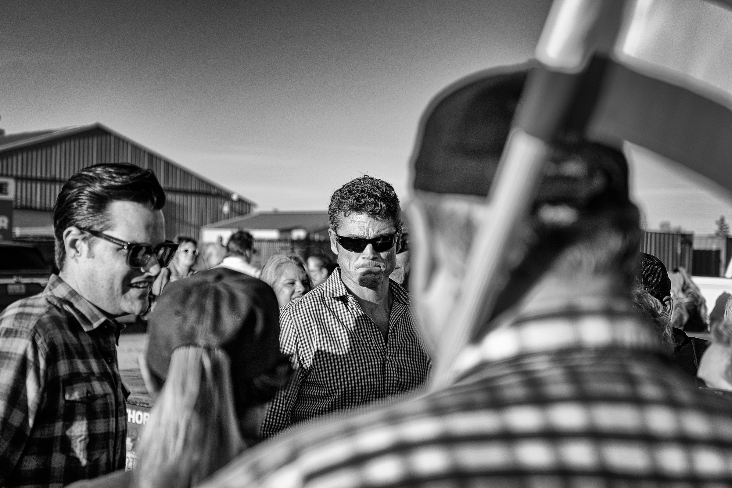 A black and white photograph of Joe Kent in a checkered shirt surrounded by supporters at an outdoor event with industrial buildings in the background. Florida congressman Matt Gaetz stands to his right as they greet two people in front of them.