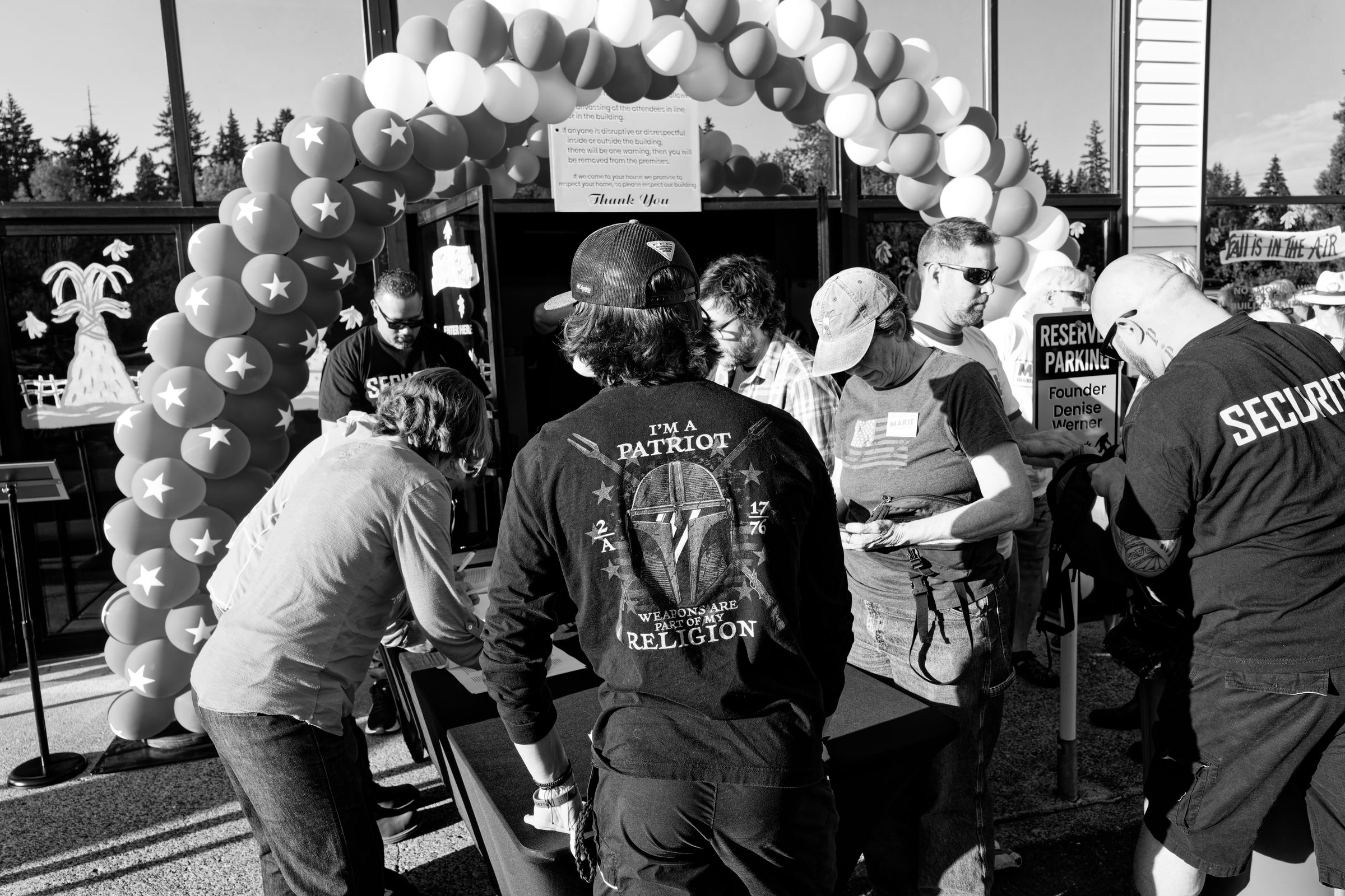 A black and white photo of a political event entrance featuring a balloon arch with stars, security personnel, and attendees. A young man in a baseball cap stands with his back to the camera; his shirt reads "I'M A PATRIOT WEAPONS ARE PART OF MY RELIGION".