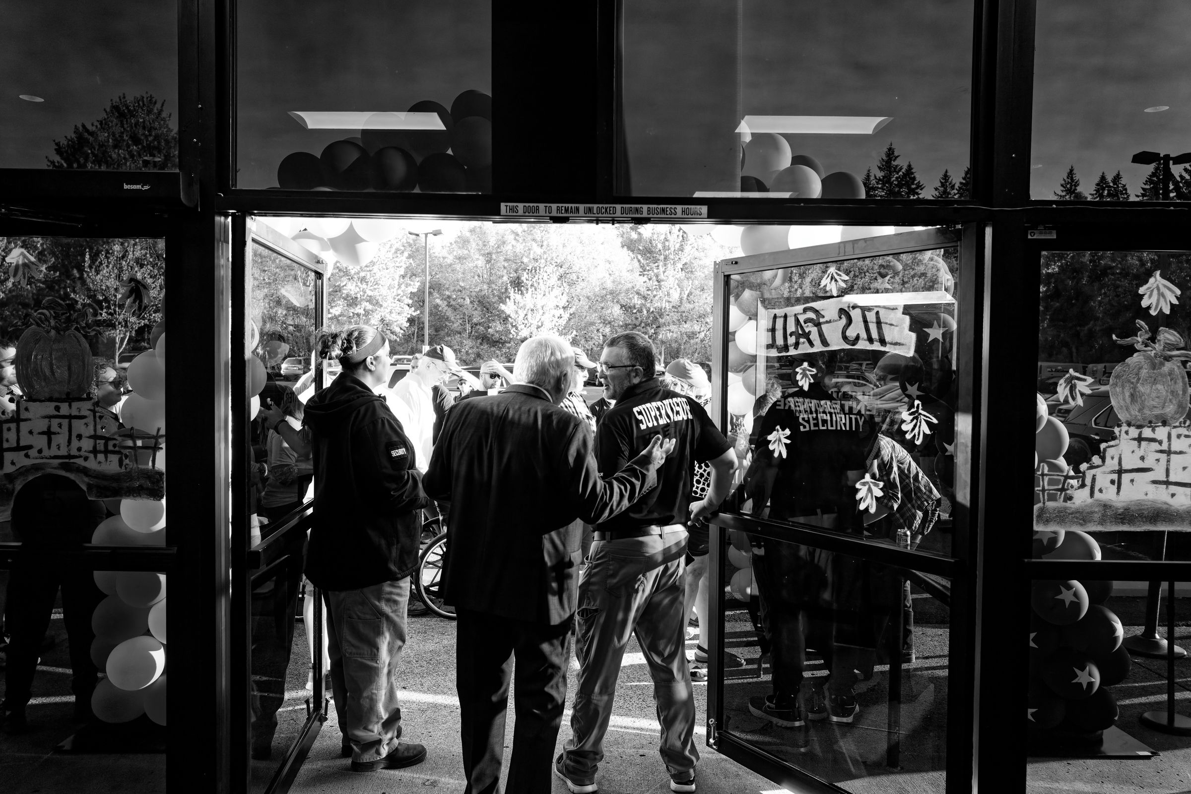 A black and white photograph of event security personnel at a venue entrance with balloons visible on the outside where attendees wait to enter.