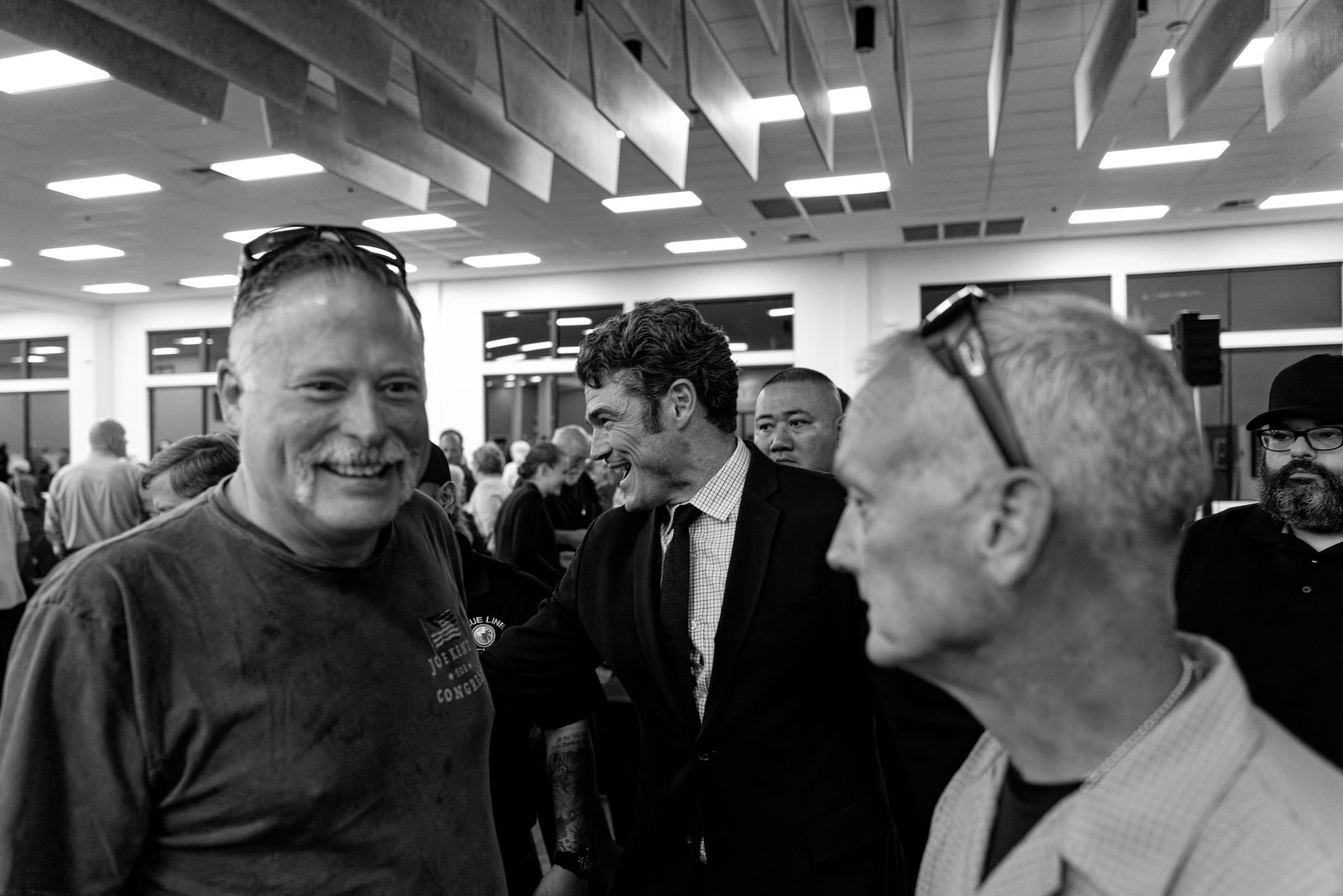 A black and white image of a political event showing a suited Joe Kent engaging with supporters in a crowded indoor room. Two men are visible in the foreground, one smiling towards the other. His shirt reads "JOE KENT FOR CONGRESS".