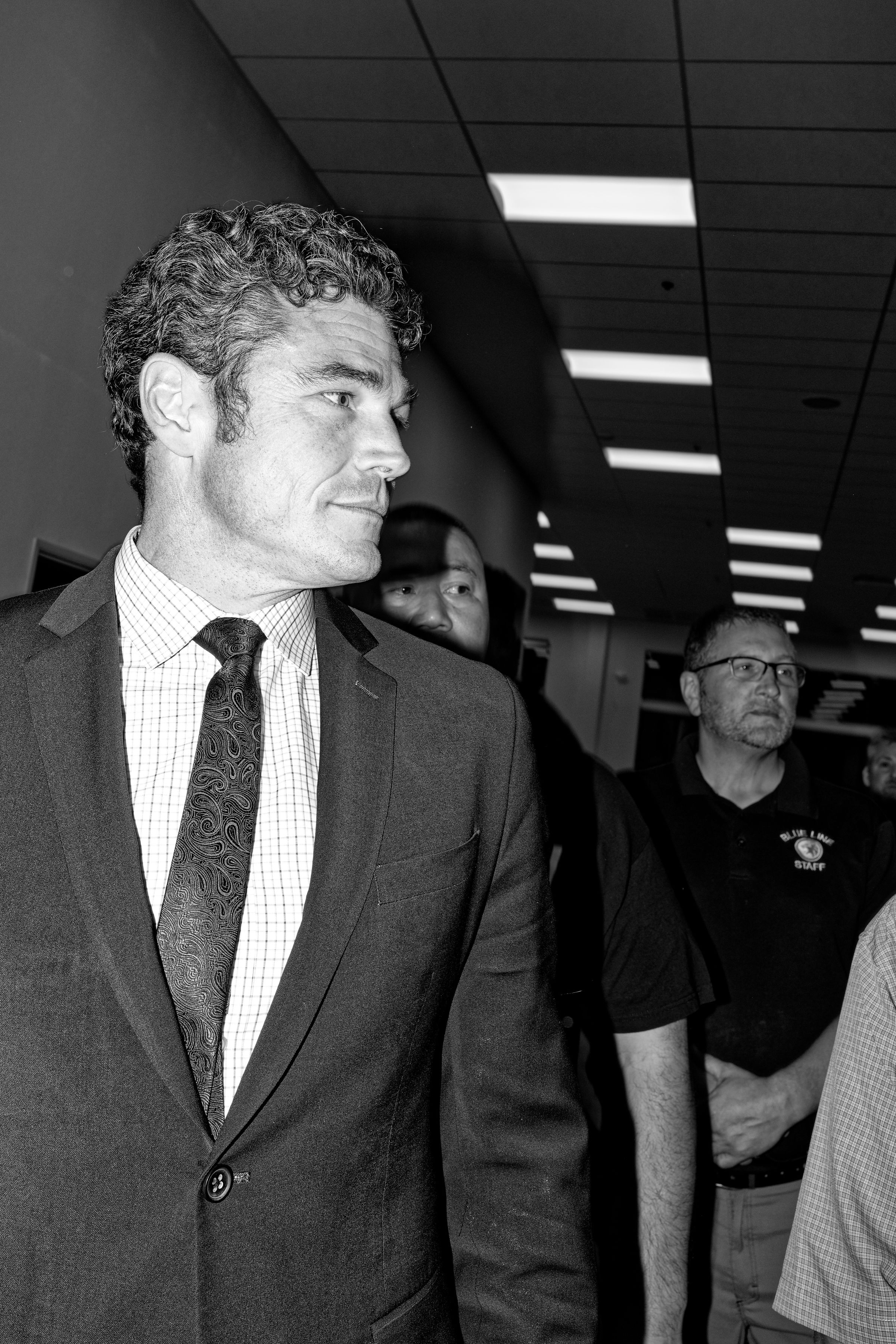 A close-up black and white photo of Joe Kent wearing a suit and tie and looking to the right of the frame. The camera's flash produces a shadow that partially obscures the face of a man over Kent's left shoulder, revealing his eyes to be looking in the same direction as Kent's. Another many stands nearby, his hands folded calmly in front of him as he observes the scene; his shirt displays an indistinct seal with the words "BLUE LINE STAFF".