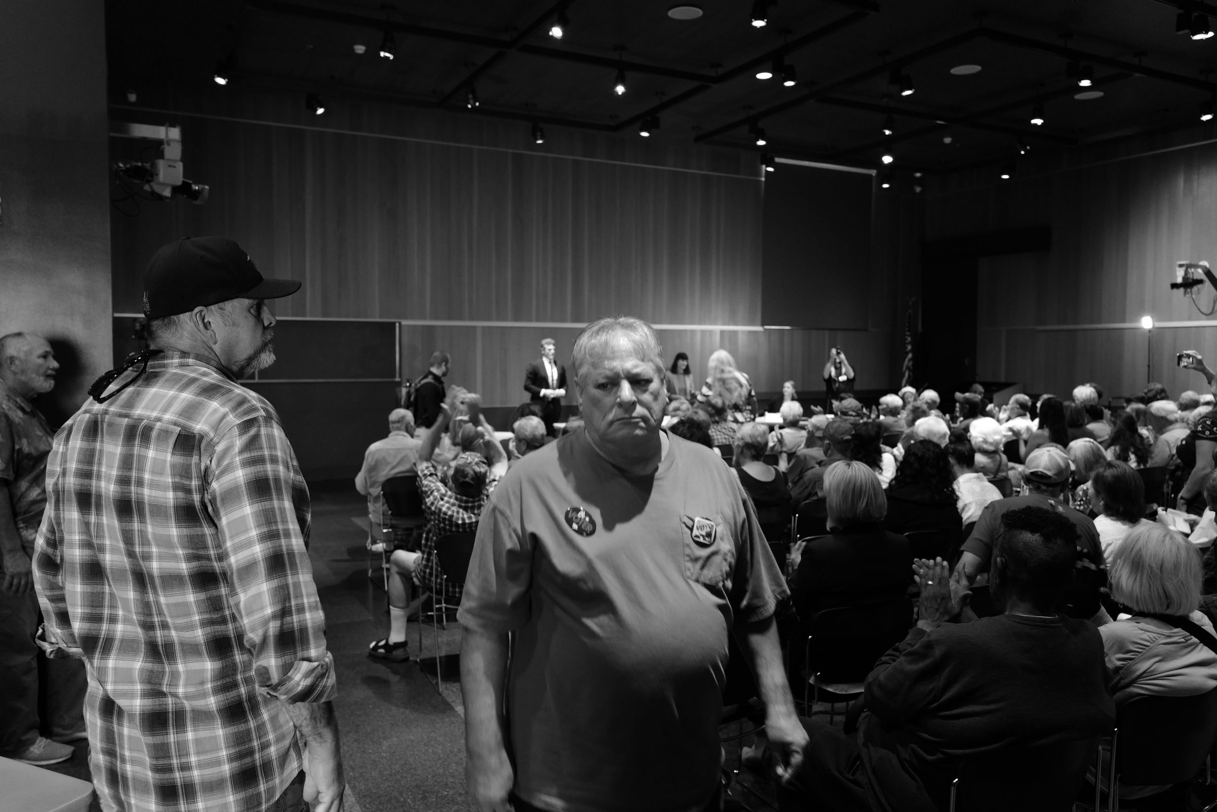 A black and white photo of an indoor campaign event. A despondent man wearing a button that reads "VOTER" moves towards the camera, leaving a crowded room of seated people behind him. A stage with a podium is visible at the front of the room. Another man in a plaid shirt and baseball cap observes the scene, partially facing the camera.