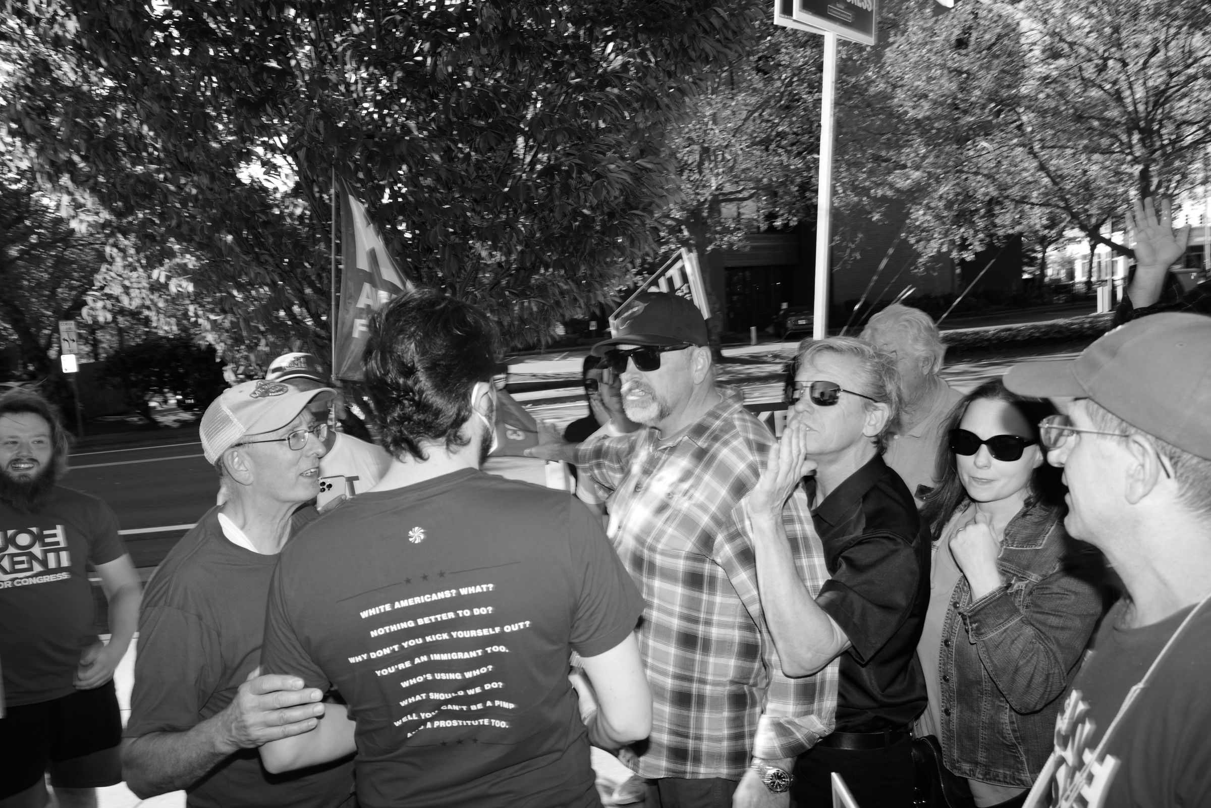 A black and white photo of an outdoor political event. A man in a plaid shirt and baseball cap stands with of a crowd of gesticulating Joe Kent supporters, many wearing campaign t-shirts and hats and holding signs. Trees line the street behind them. The Kent supporters surround a man wearing a face mask, his back to the camera. The back of his shirt reads, "White Americans? What? Nothing better to do? Why don't you kick yourself out? You're an immigrant too." — a quote from a lyric by The White Stripes. Another man in a baseball cap holds his arm while speaking imploringly to him.