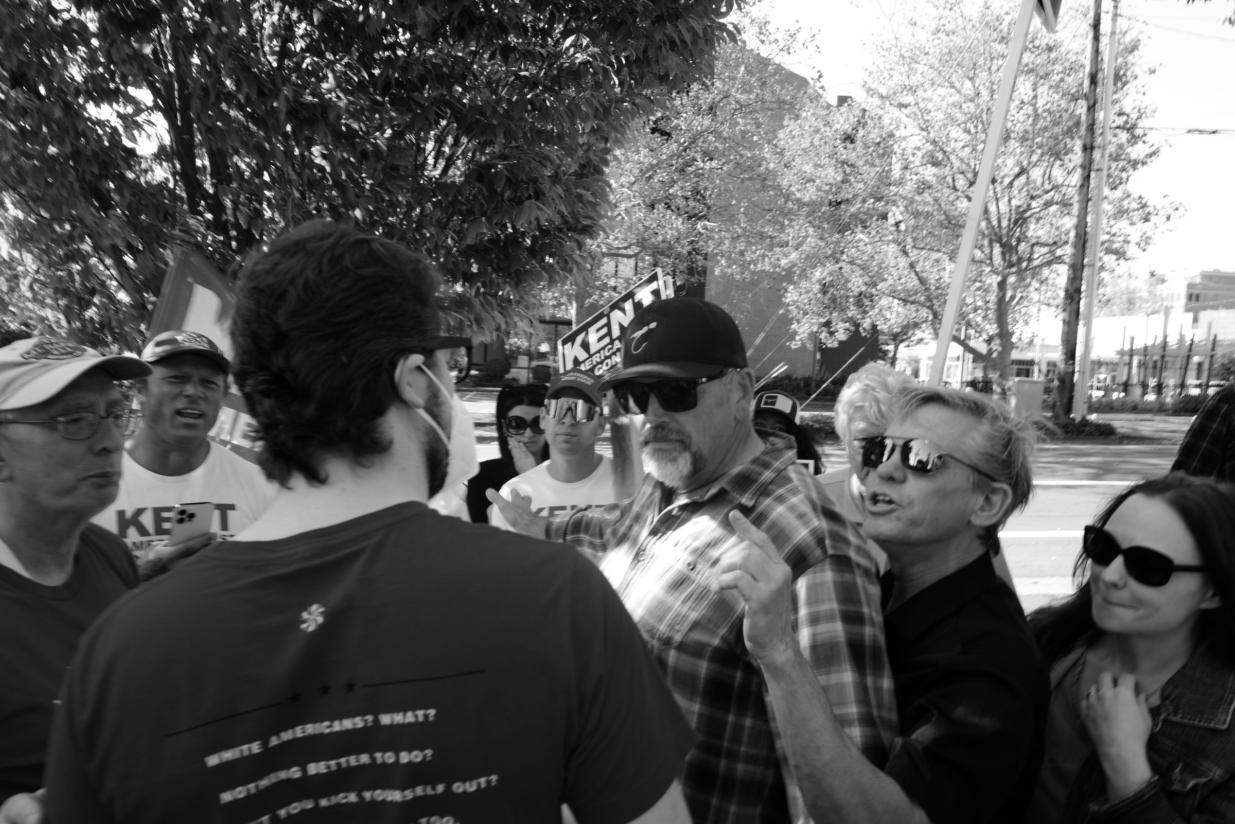 A black and white photo of an outdoor political event. A man in a plaid shirt and baseball cap stands with of a crowd of jeering Joe Kent supporters arrayed behind him, some wearing campaign t-shirts and hats and holding signs. Trees line the street behind them all. The Kent supporters face a lone man wearing a face mask, his back to the camera. The back of his shirt reads, "White Americans? What? Nothing better to do? Why don't you kick yourself out?" — a quote from a lyric by The White Stripes that ends with, "You're an immigrant too."