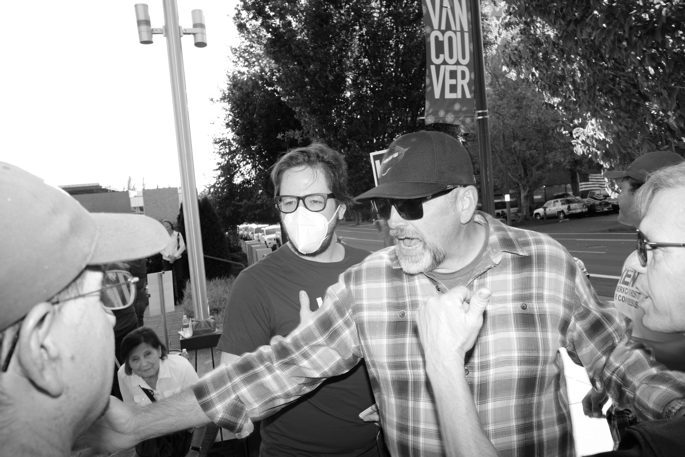 A black and white photo of an outdoor political event. A man in a plaid shirt and baseball cap gestures emphatically while speaking to several other men off-frame. Behind him, a person wearing a face mask is visible. A street banner with the letters "VAN COU VER" is partially visible in the background.