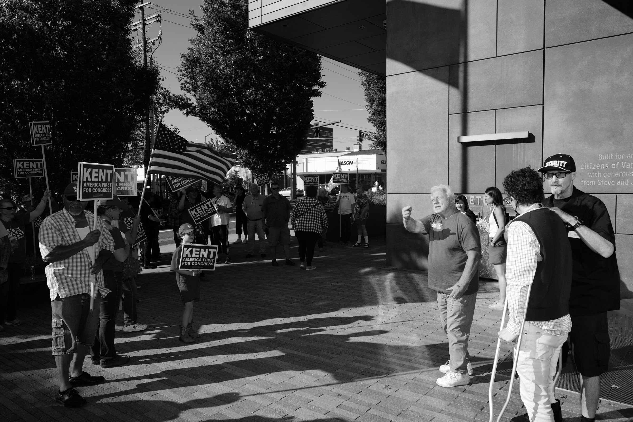 A black and white photo of two groups of people facing off across a paved walkway outside a tall angular building. Joe Kent supporters hold numerous campaign signs and American flags on one side, jeering at a much smaller group opposite from them. An older man with gray hair stands out from the opposing crowd, gesturing with his middle finger at the Kent supporters; this man was called a "Nazi" by Kent supporters because of his support for Ukraine.