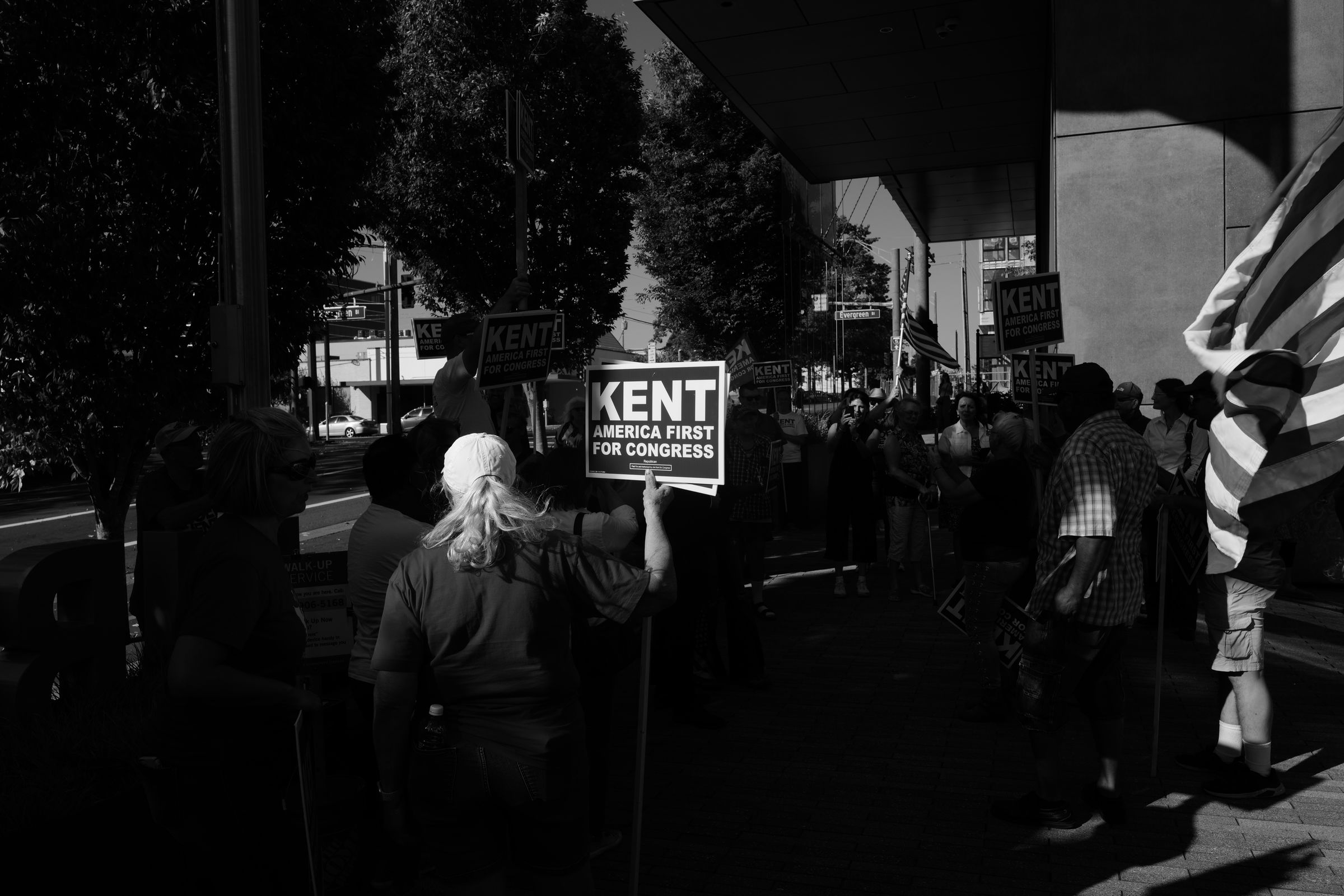 A black and white photo of Joe Kent supporters holding campaign signs outside the overhung entrance to a tall building. The shadows from the architecture contrast with the afternoon sunlight, illuminating a "KENT AMERICA FIRST FOR CONGRESS" sign in the center of the image.