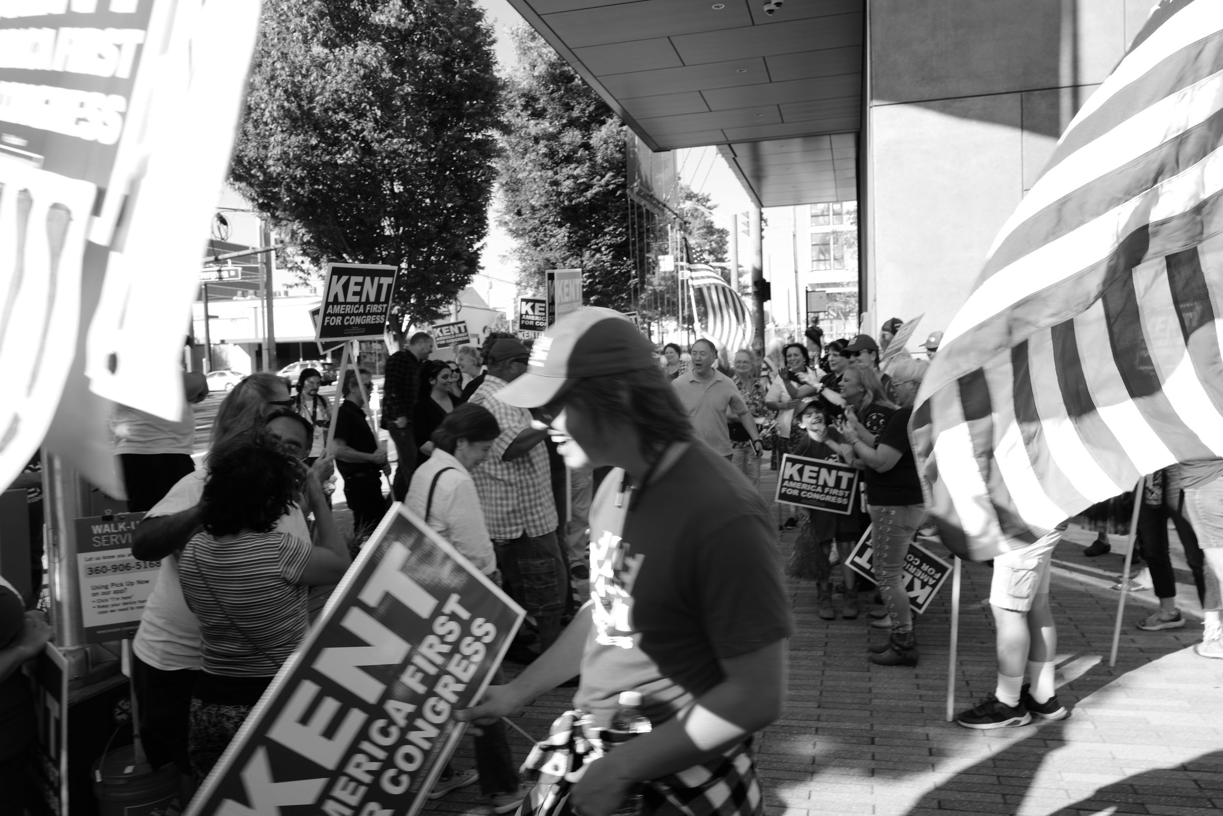 A black and white photo of a dense crowd of Joe Kent supporters holding "KENT AMERICA FIRST FOR CONGRESS" signs and American flags. A young man holding a campaign sign is caught in motion at the center of the frame, laughing smugly.