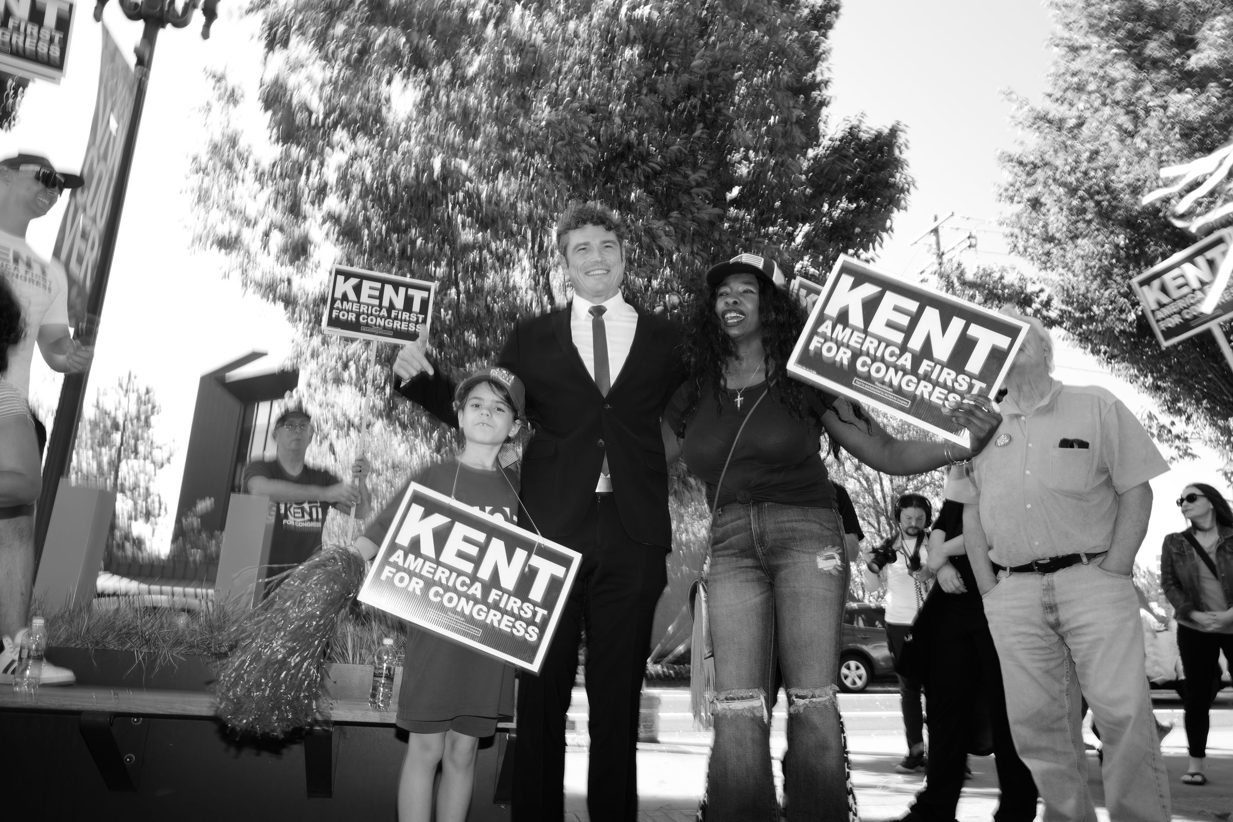 A black and white image of Joe Kent wearing a suit and tie, standing between a young boy and a woman, all holding "KENT AMERICA FIRST FOR CONGRESS" signs. Kent is smiling broadly. The scene unfolds at an outdoor campaign event with trees in the background and other supporters visible.