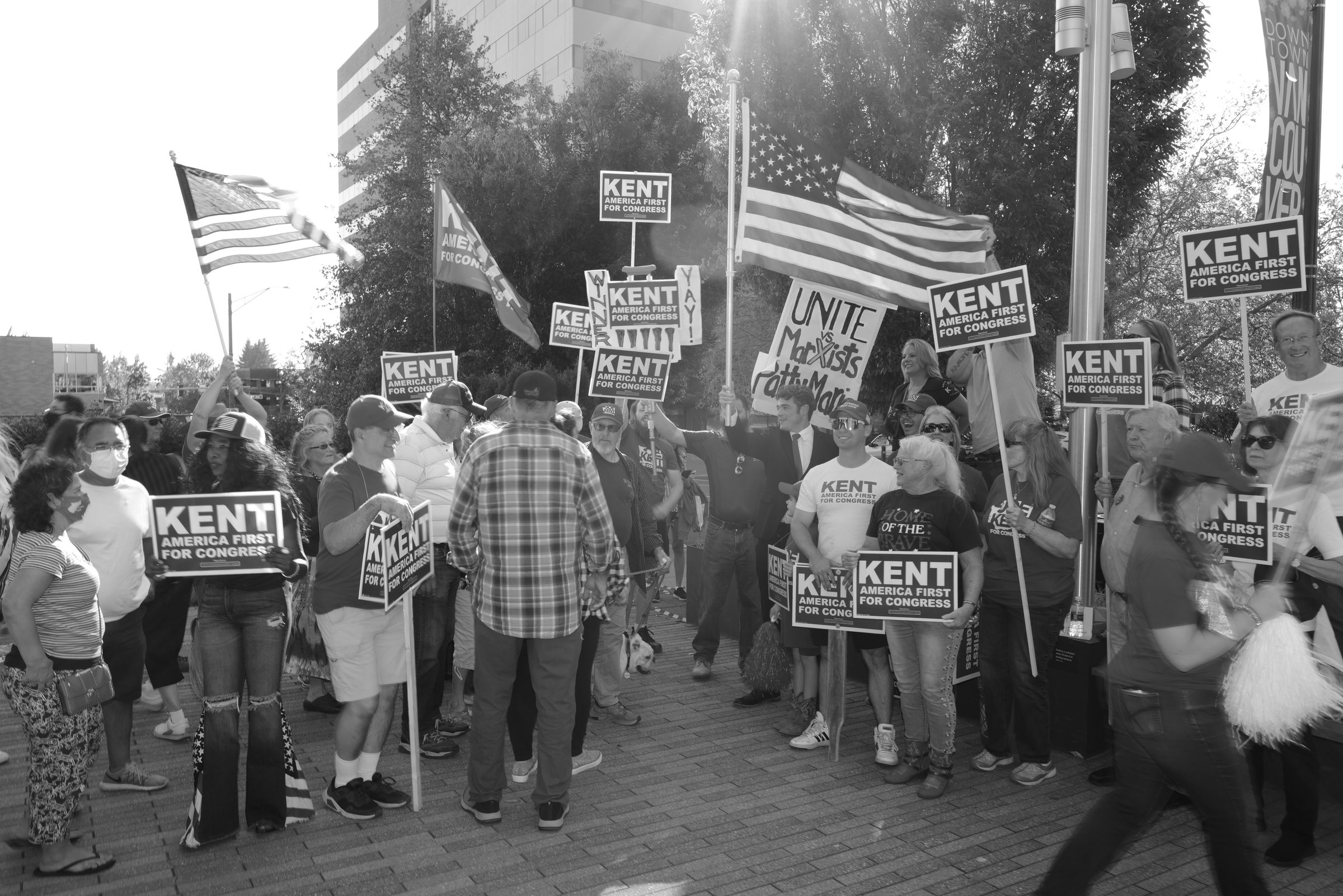 A black and white photograph of a crowded political rally for Joe Kent's congressional campaign. Supporters hold numerous "KENT AMERICA FIRST FOR CONGRESS" signs. American flags wave prominently, including a large one in the center. The scene is outdoors with trees and buildings visible. Supporters of various ages are present, some wearing Kent campaign t-shirts. A few people wear face masks, suggesting this is during the COVID-19 pandemic.